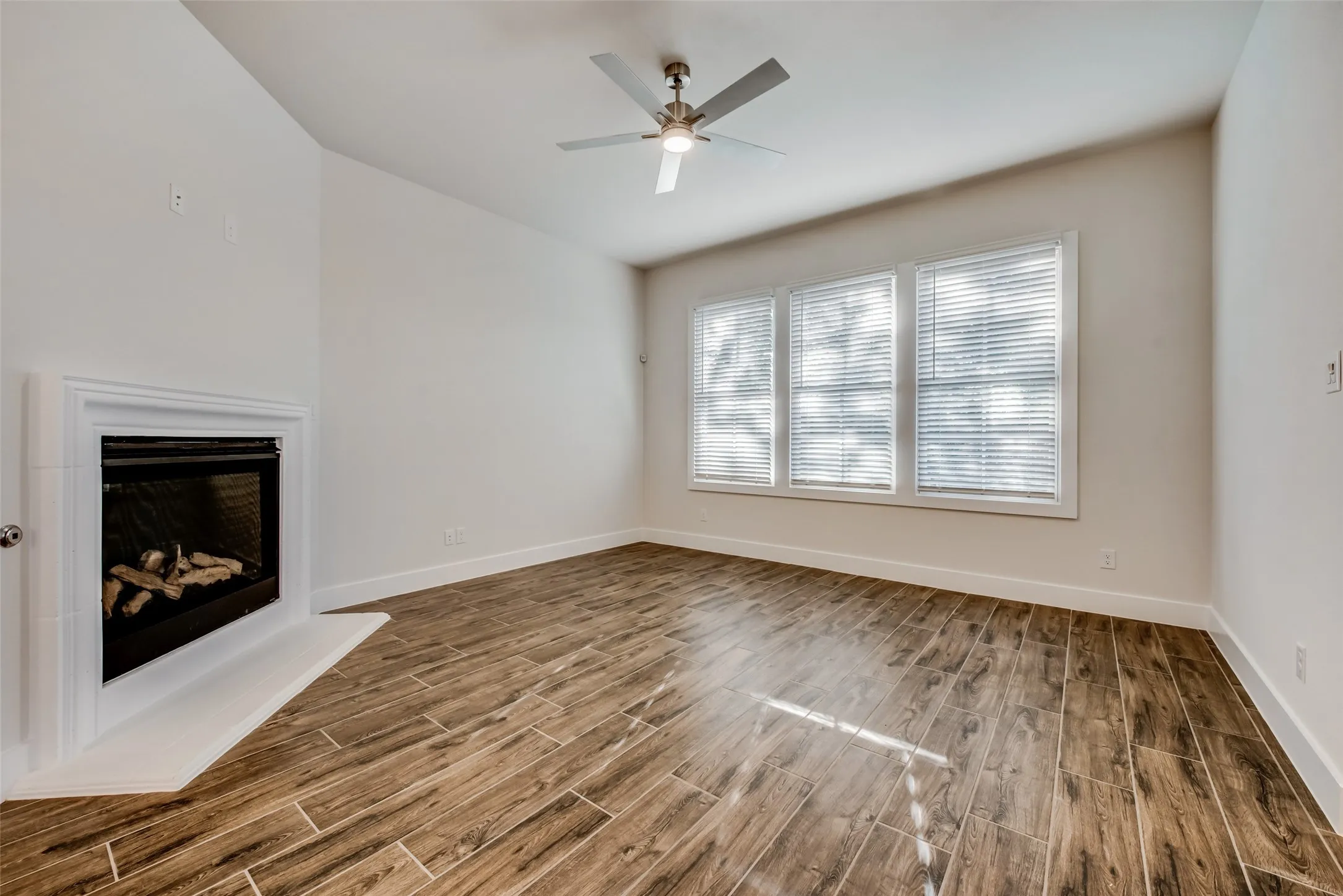 Spacious room featuring wood-finish tile flooring, a corner fireplace with a white mantel, and three windows with horizontal blinds