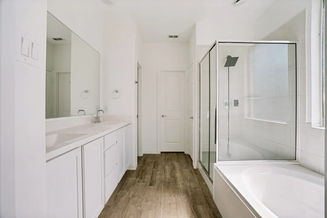 Dual vanity with white cabinetry, wood-finish flooring, a frameless glass shower enclosure, a built-in soaking tub, and contemporary fixtures