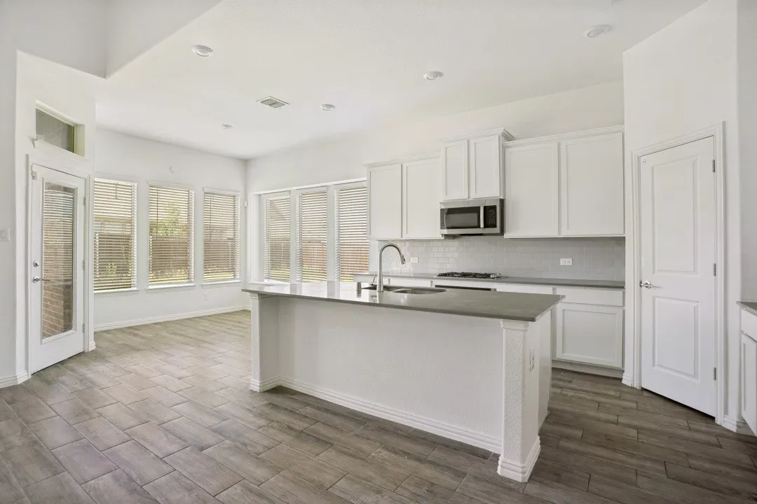 Open-concept kitchen featuring an island with a sink, stainless steel microwave, white shaker cabinetry, and wood-finish tile flooring