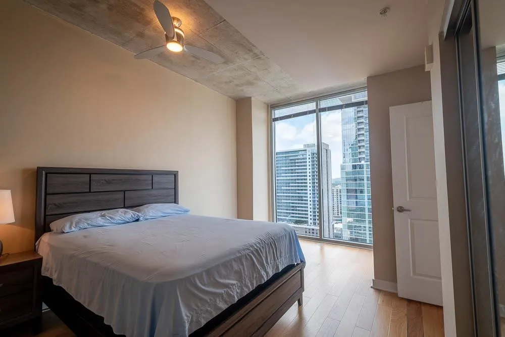 Floor-to-ceiling windows with urban skyscraper backdrop, hardwood flooring, and a modern ceiling fan with integrated lighting