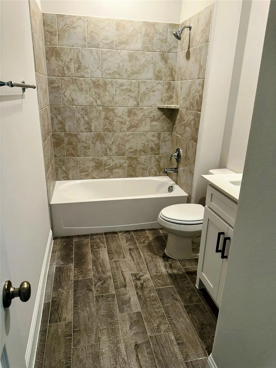 Bathroom featuring wood-finish flooring, a white vanity with black hardware, a white toilet, and a bathtub with tiled shower surround