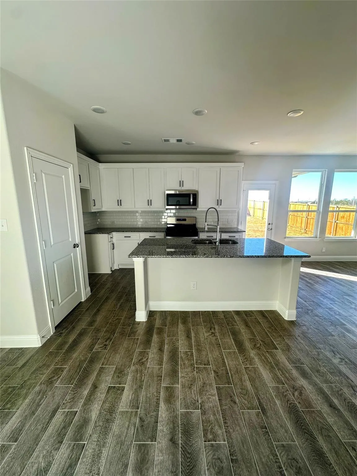 Open-concept kitchen featuring wood-finish flooring, a central island with a sink, white cabinetry with dark hardware, stainless steel appliances, and a subway tile backsplash