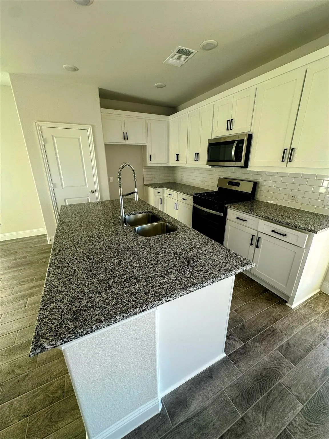 Kitchen featuring a central island with a double sink and granite countertop, white shaker-style cabinetry, stainless steel appliances including a microwave and range, a subway tile backsplash, and wood-finish tile flooring
