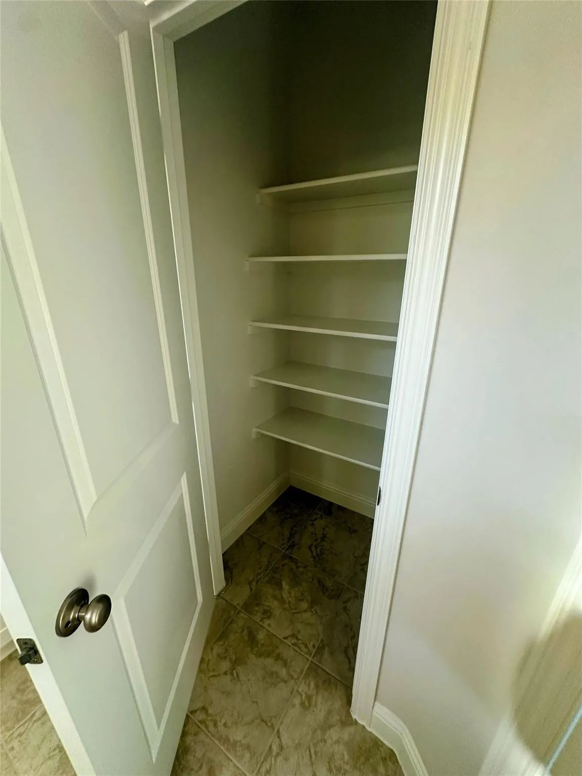 Recessed pantry with five levels of shelving, neutral-toned tile flooring, and a white panel door with a bronze-finish knob