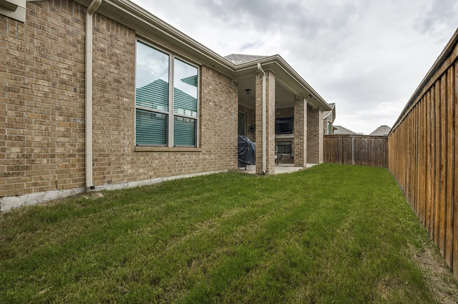 Grassed side yard featuring a privacy fence, brick exterior, single-hung windows, and a covered patio with brick columns