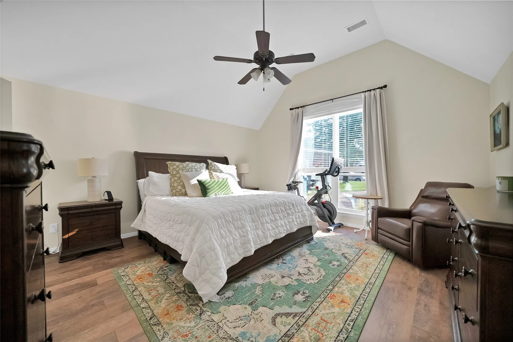Spacious room featuring vaulted ceilings, wood-finish flooring, a ceiling fan with integrated lighting, and ample natural light from a large window with blinds and drapes