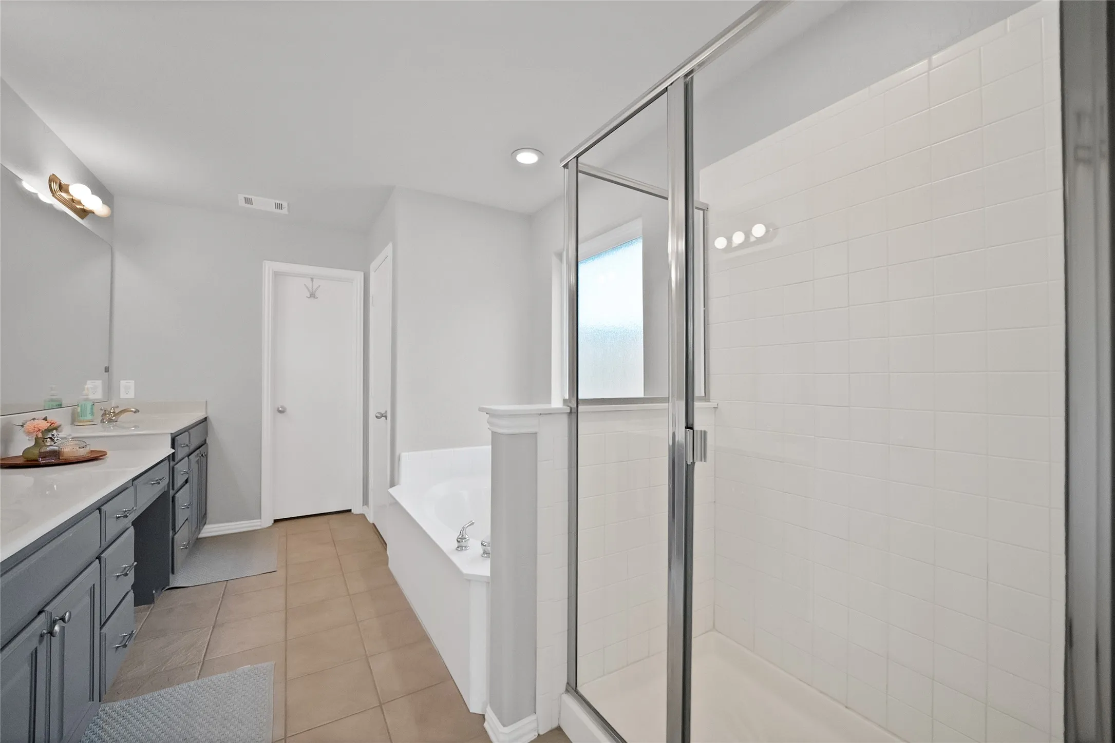 Bathroom with a glass-enclosed shower, a built-in soaking tub, and a double vanity featuring gray cabinetry and light-toned countertops