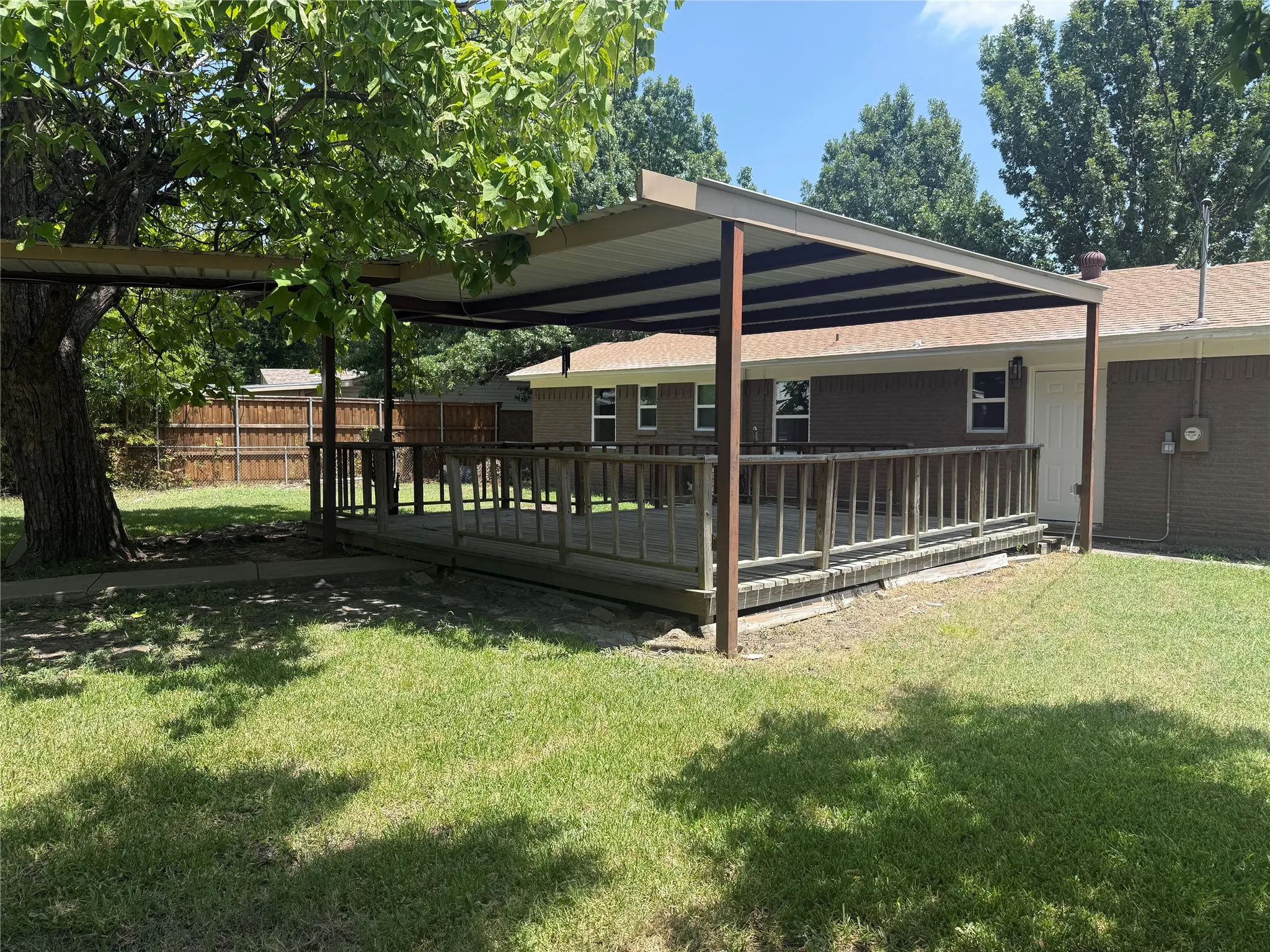 Spacious backyard featuring a covered wood deck with railing, a grass lawn, and mature trees providing shade