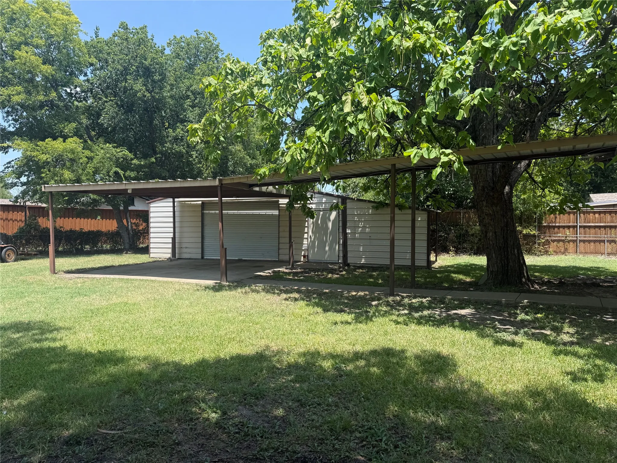 Expansive backyard featuring a metal carport structure, a detached storage shed with a roll-up door, a mature shade tree, and wood panel fencing