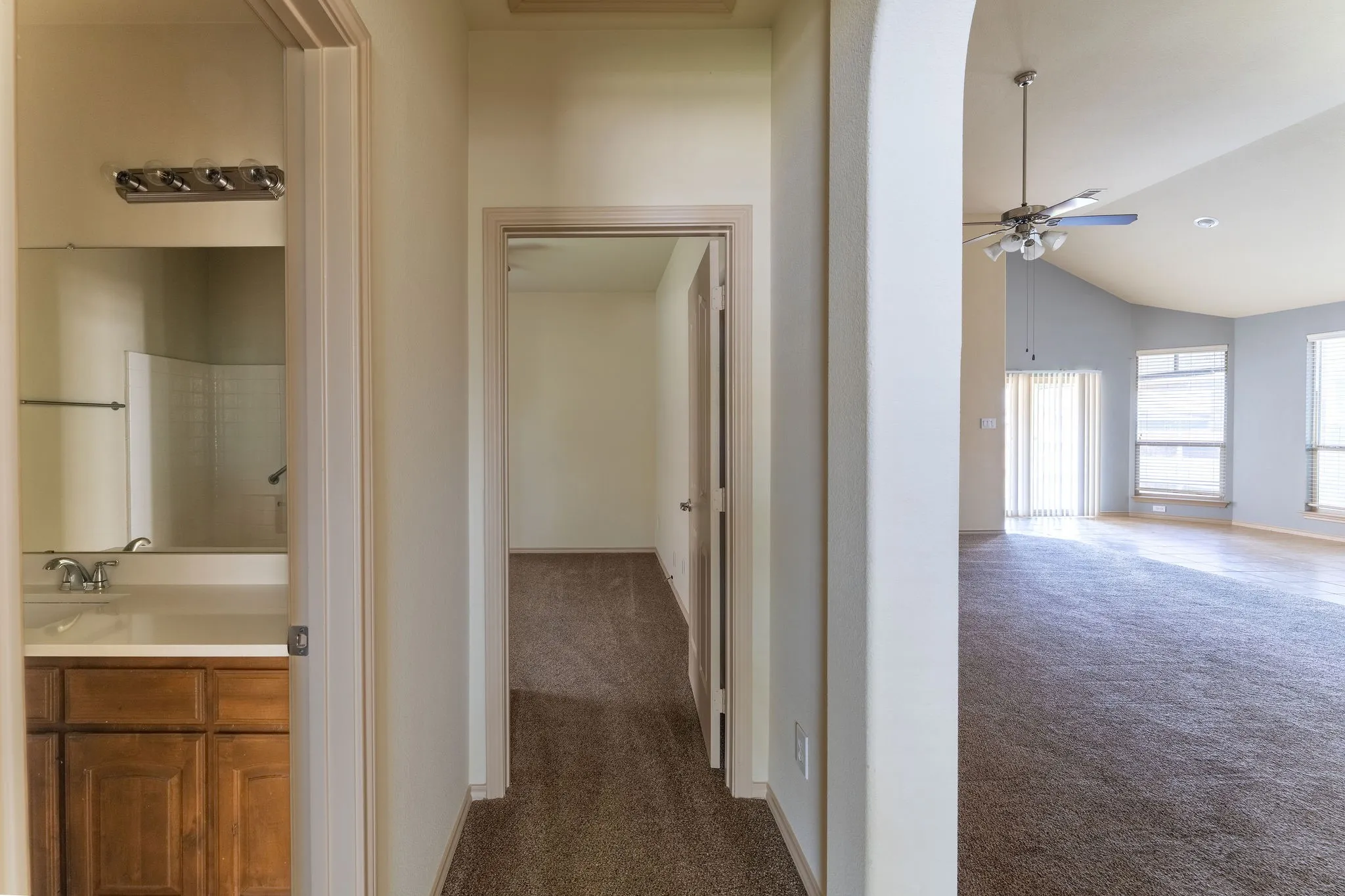 Hallway featuring textured carpet flooring, leading to a bathroom with a wood-finish vanity and a separate room