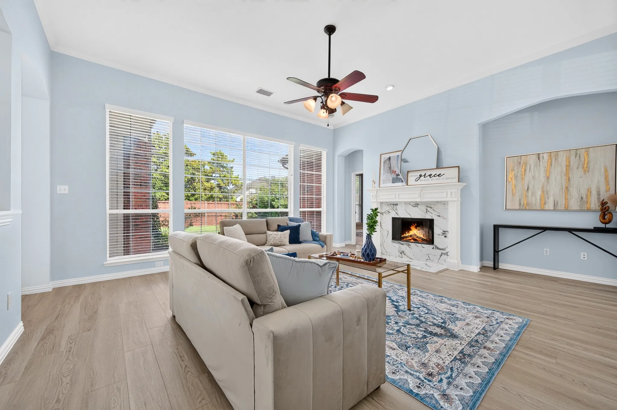 Spacious living area featuring light wood-finish flooring, a white mantel fireplace with a stone-look surround, an arched entryway, and large windows with blinds