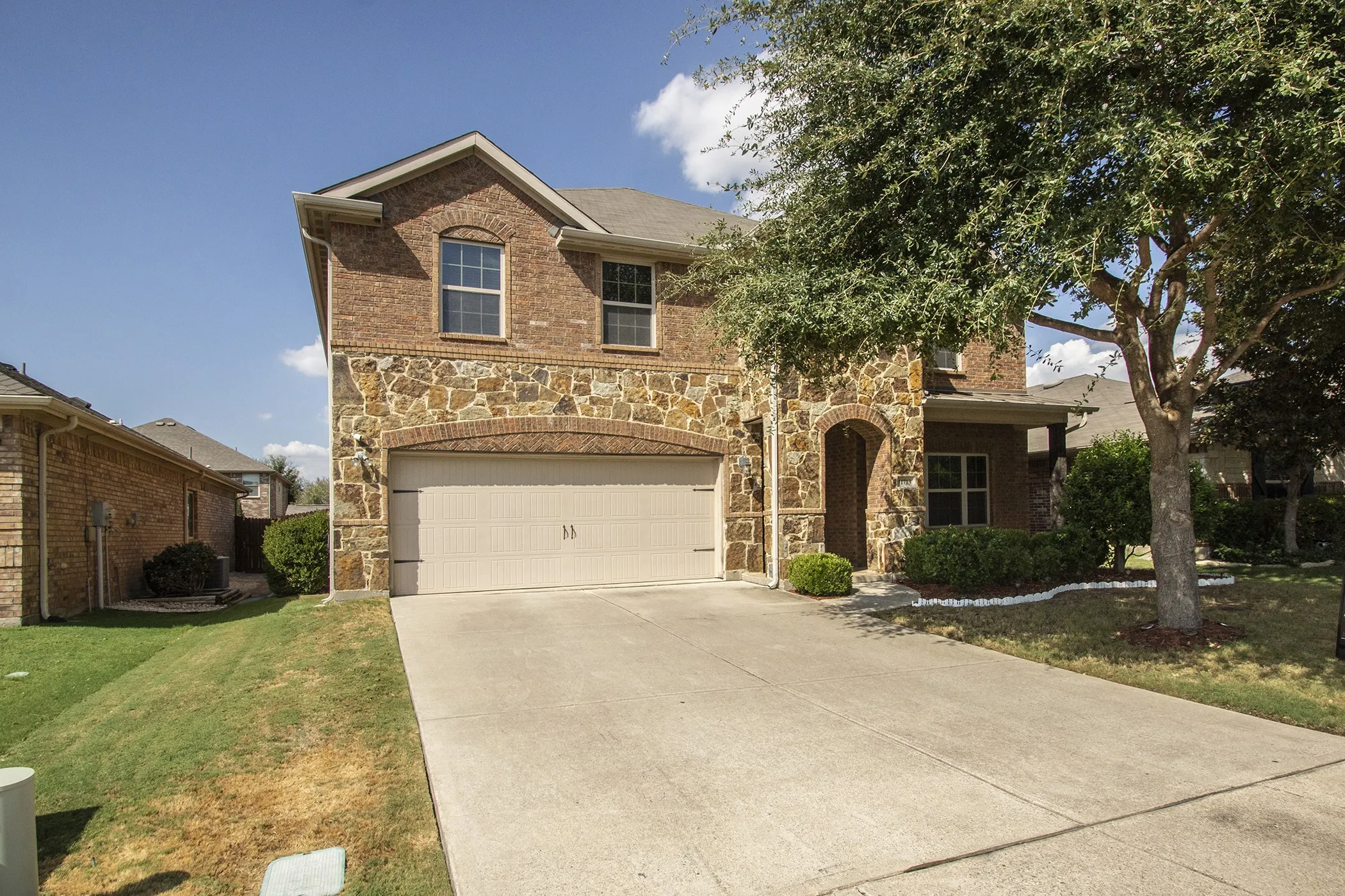 Brick and stone facade with an integrated two-car garage, arched entry, concrete driveway, and mature landscaping
