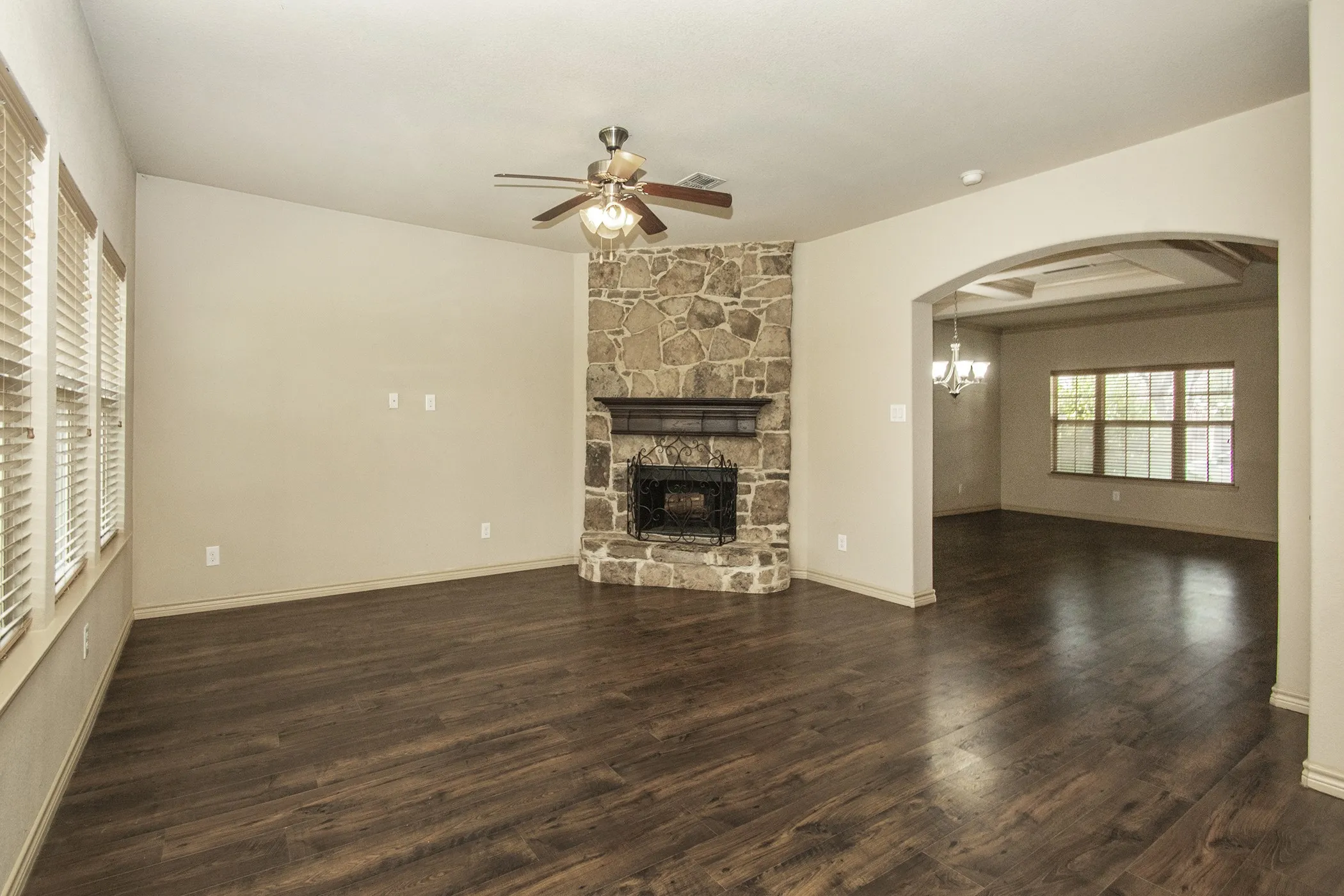 Wood-finish flooring, stone fireplace with a dark wood mantel, ceiling fan with integrated lighting, multiple windows, and an arched entryway