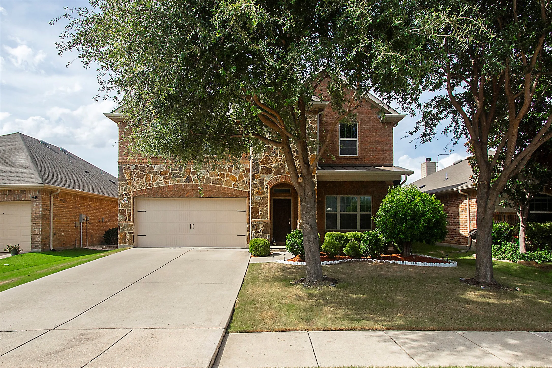 Brick and stone facade with an integrated two-car garage, arched entry, concrete driveway, and mature landscaping