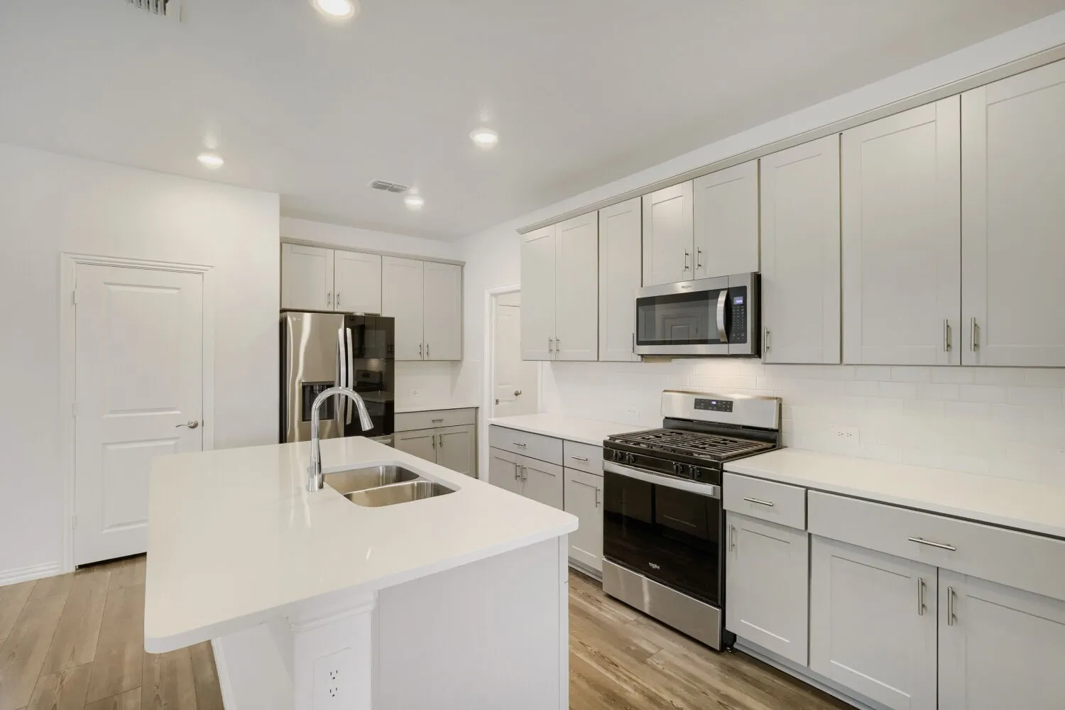 Spacious kitchen featuring a central island with a dual-basin sink, light-toned cabinetry, stainless steel appliances, white countertops, and wood-finish flooring