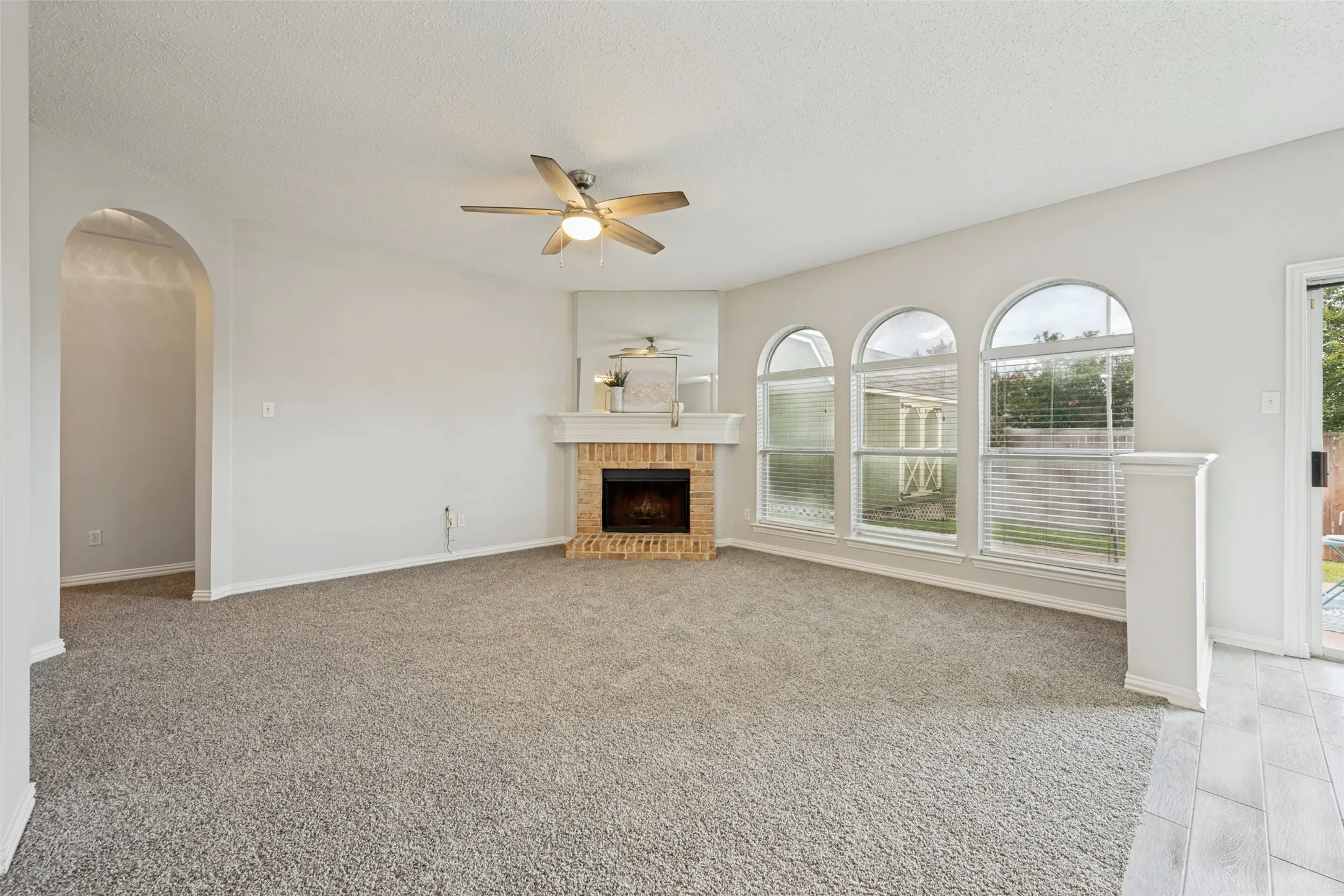 Carpeted living area featuring a corner fireplace with brick surround and white mantle, three arched windows with blinds, an arched doorway, and a ceiling fan