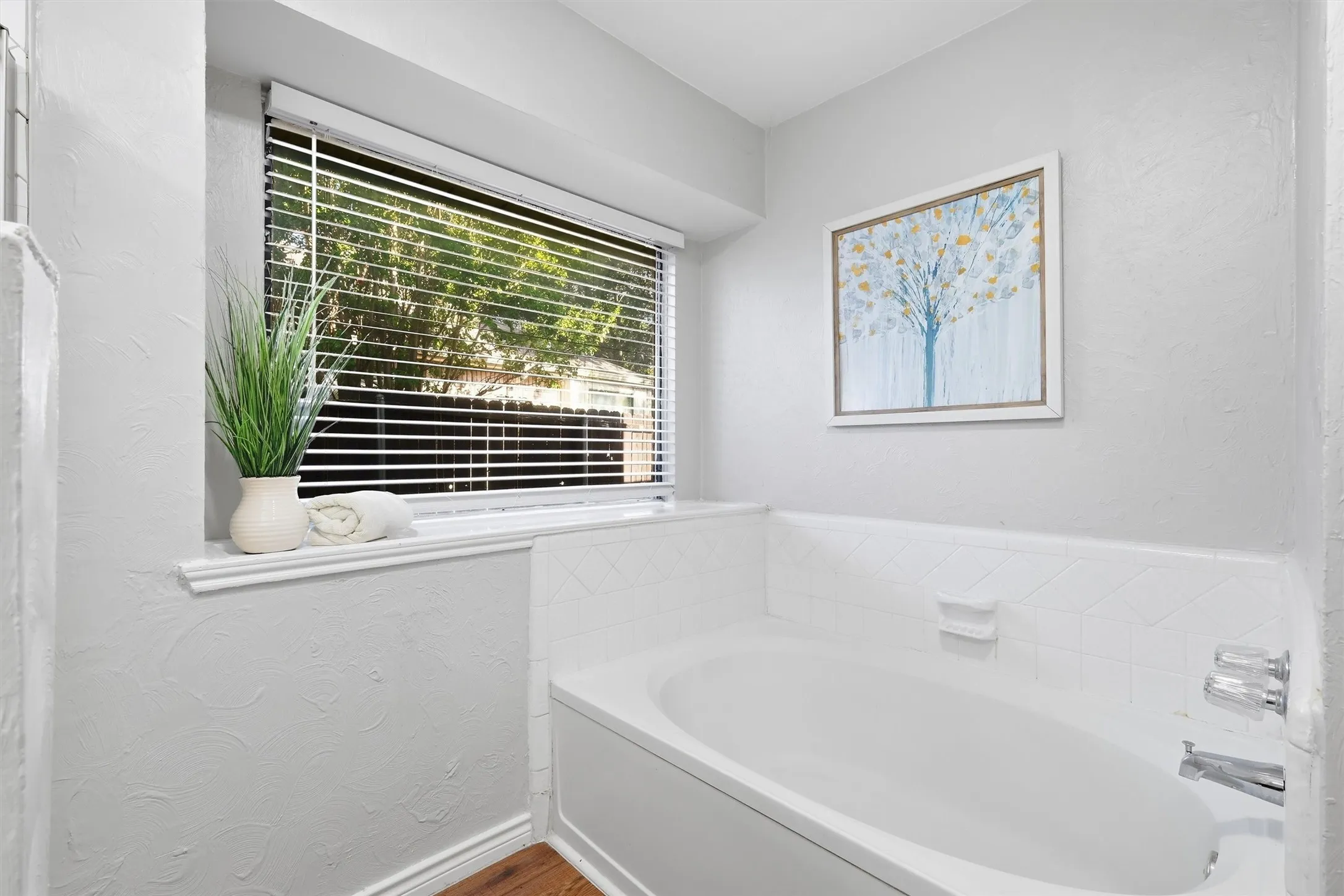 White soaking tub with chrome fixtures, a tiled surround, and a window featuring horizontal blinds