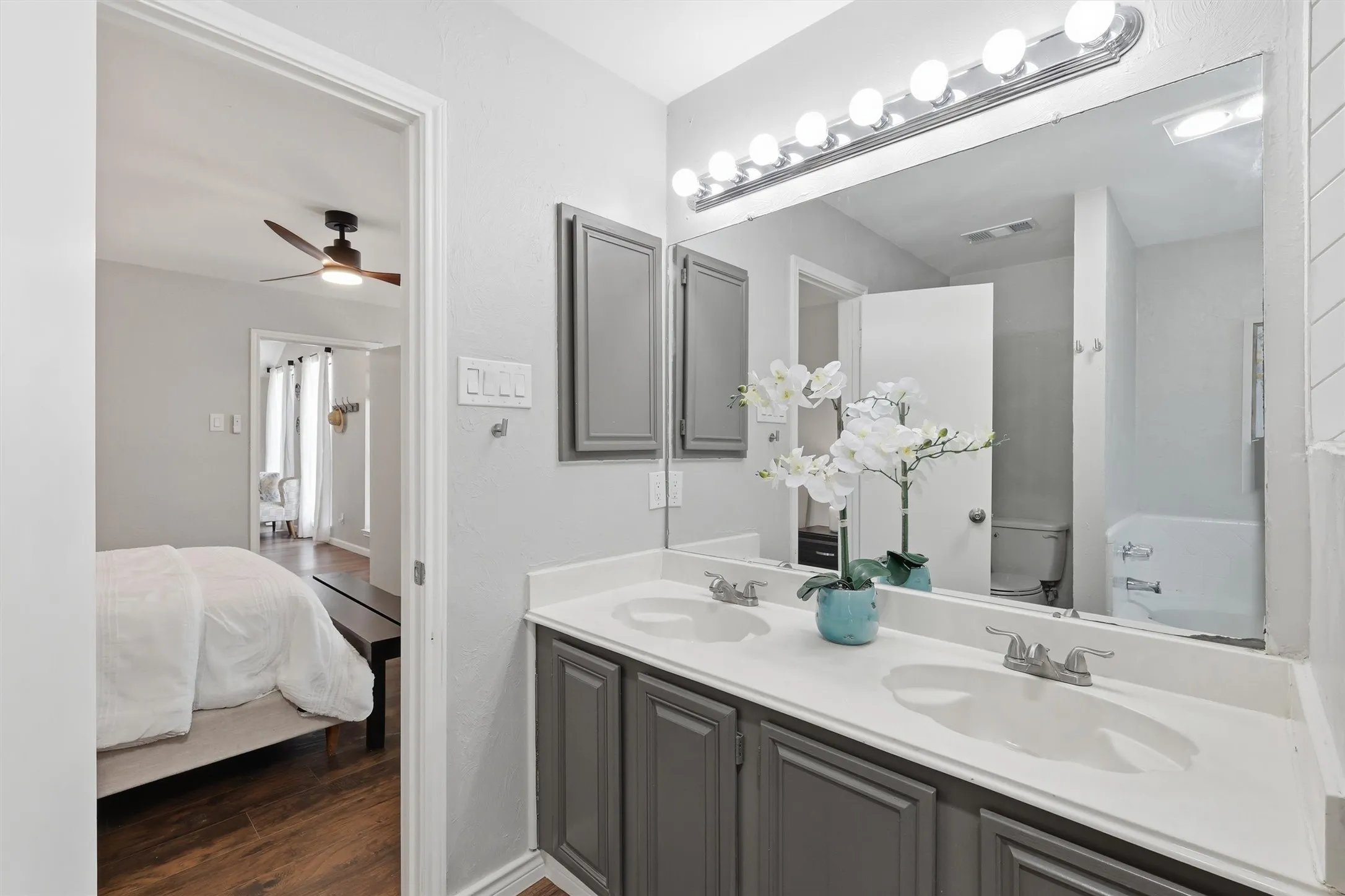 Bathroom featuring a dual vanity with a white countertop and gray cabinetry, a large vanity mirror with strip lighting, and a white bathtub with a shower