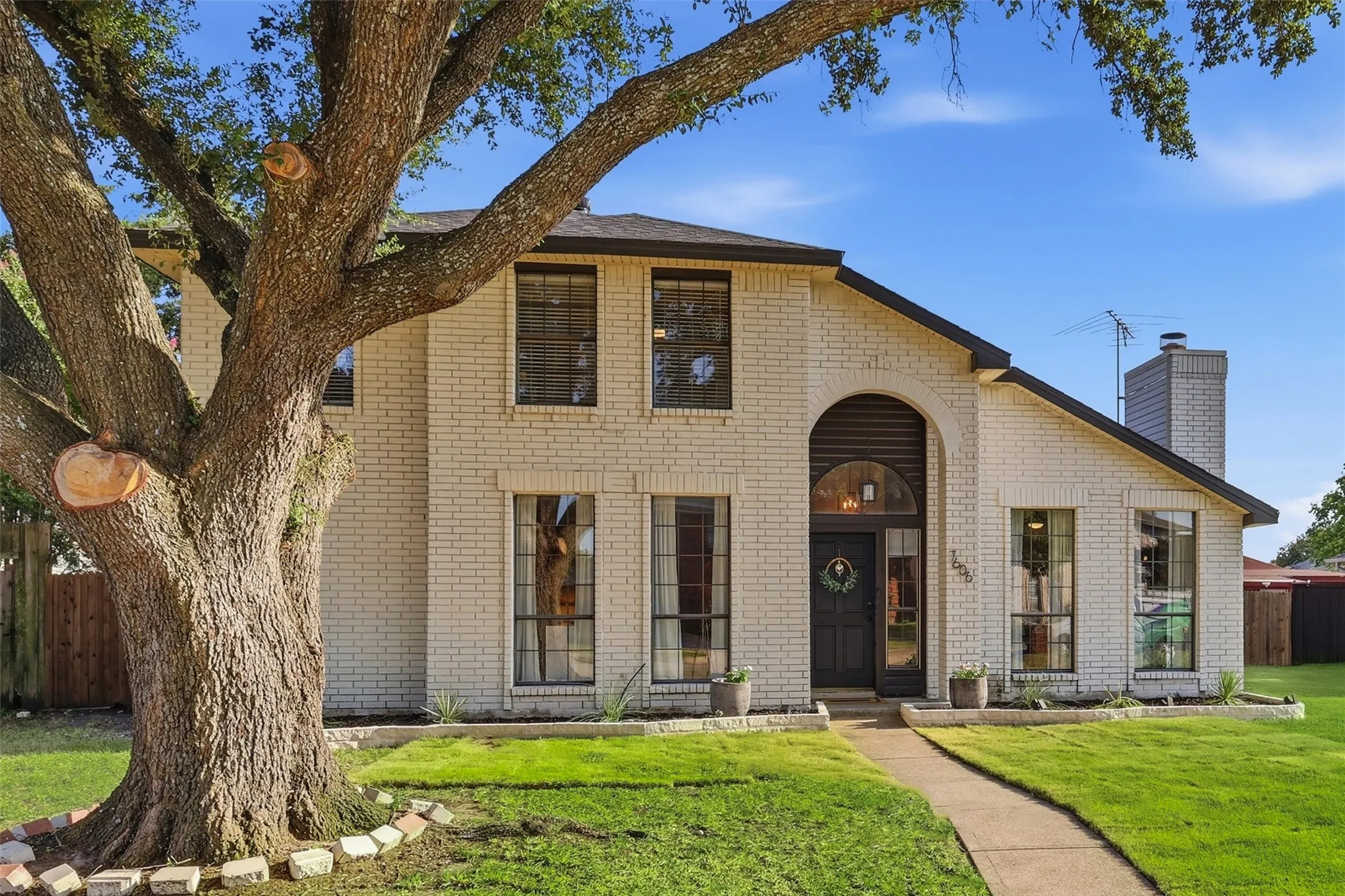 Two-story brick facade residence featuring an arched entry, a large mature tree, a paved walkway, and a landscaped front yard