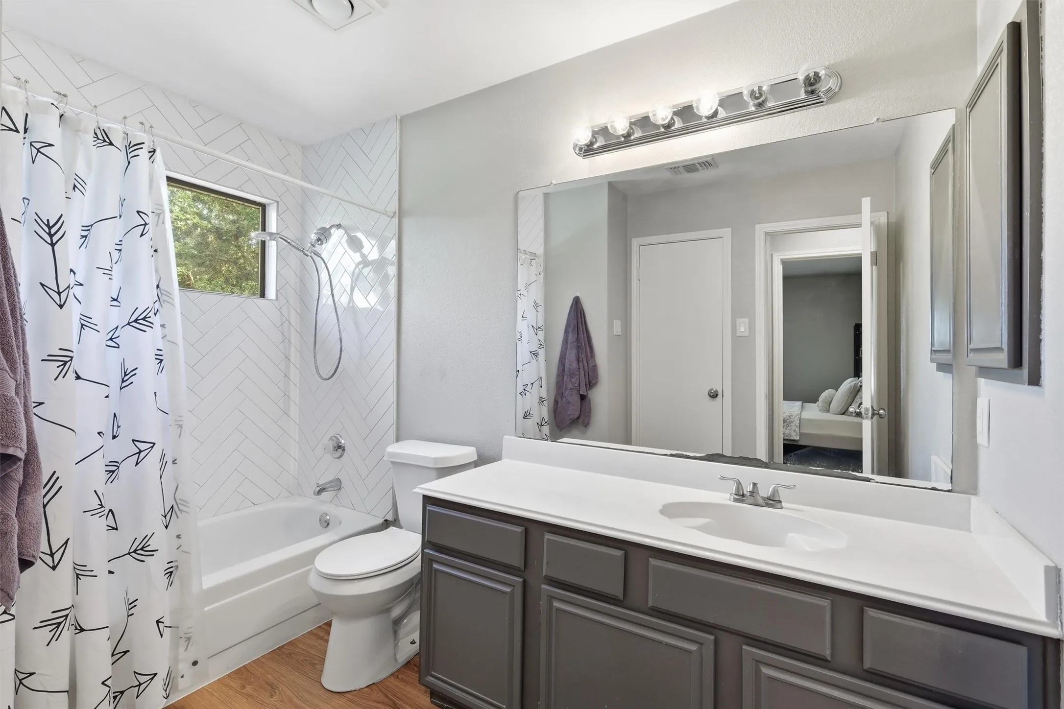Full bathroom featuring a single vanity with a white countertop and dark gray cabinetry, a rectangular mirror, a chrome light fixture, a bathtub with a shower, and white subway tile in a herringbone pattern