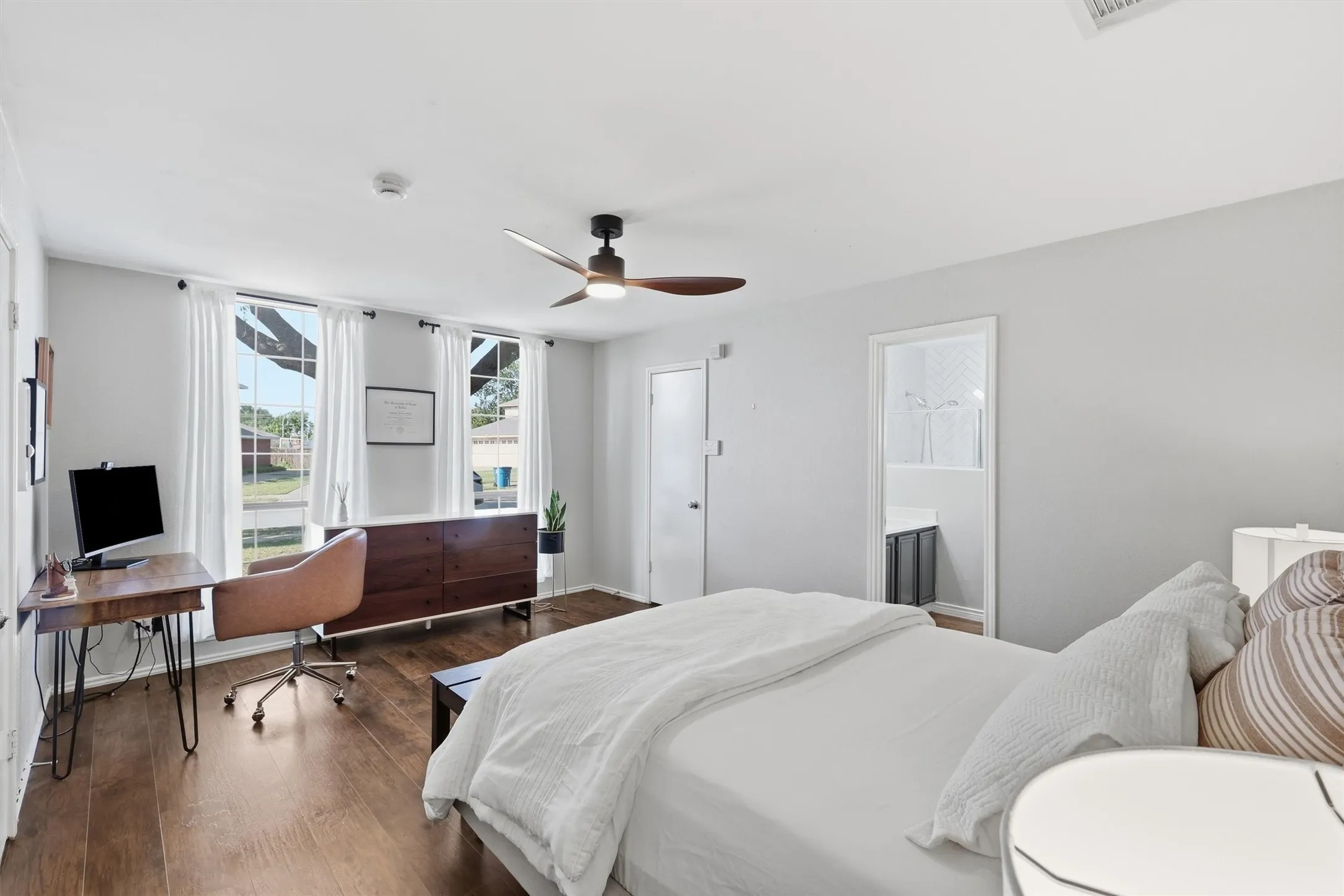 Wood-finish flooring throughout, featuring a contemporary ceiling fan, two windows with white curtains, and a doorway leading to an adjoining space