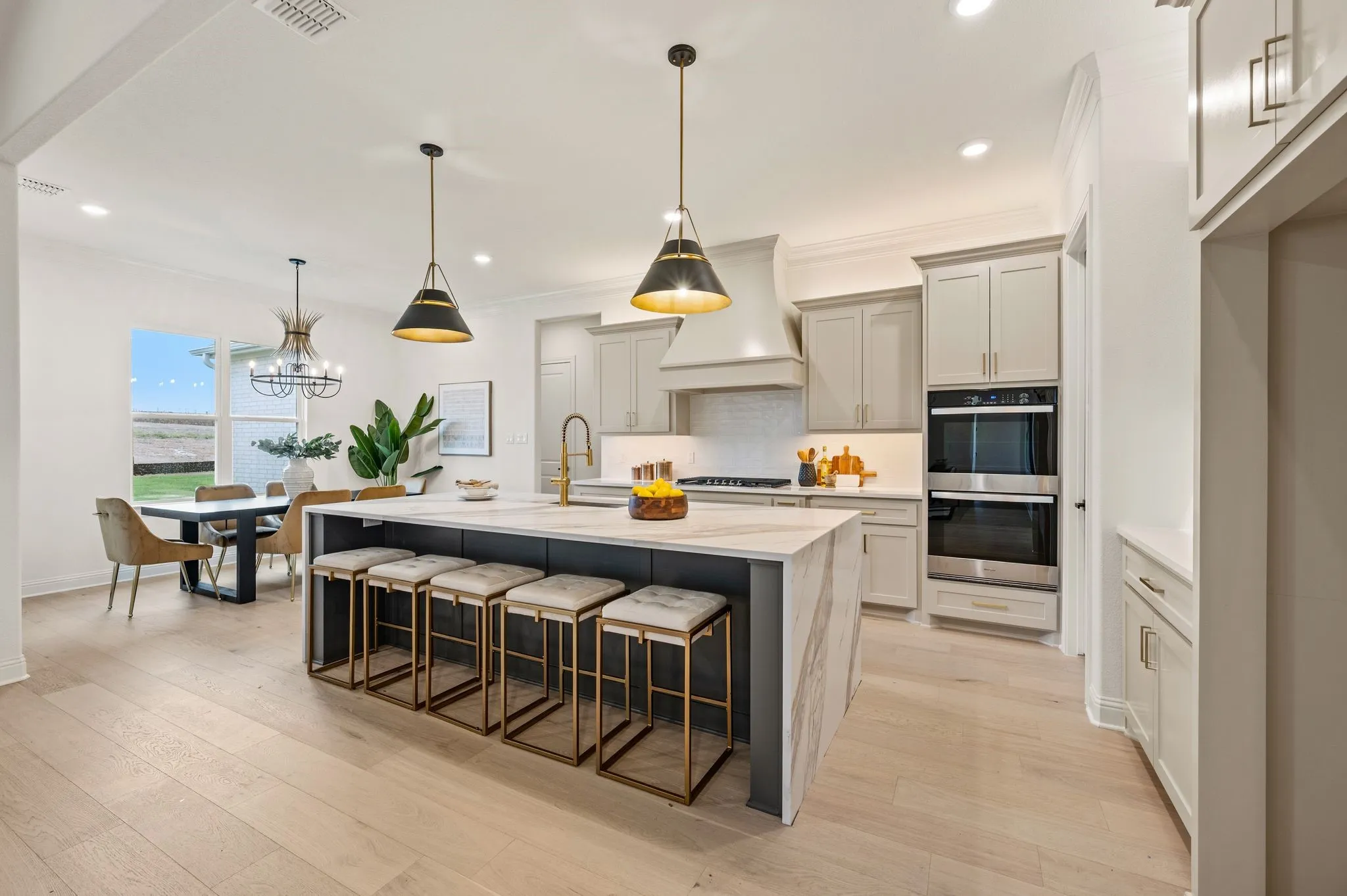 Contemporary kitchen featuring a large central island with waterfall countertop, light wood-finish flooring, light gray cabinetry, and built-in double ovens