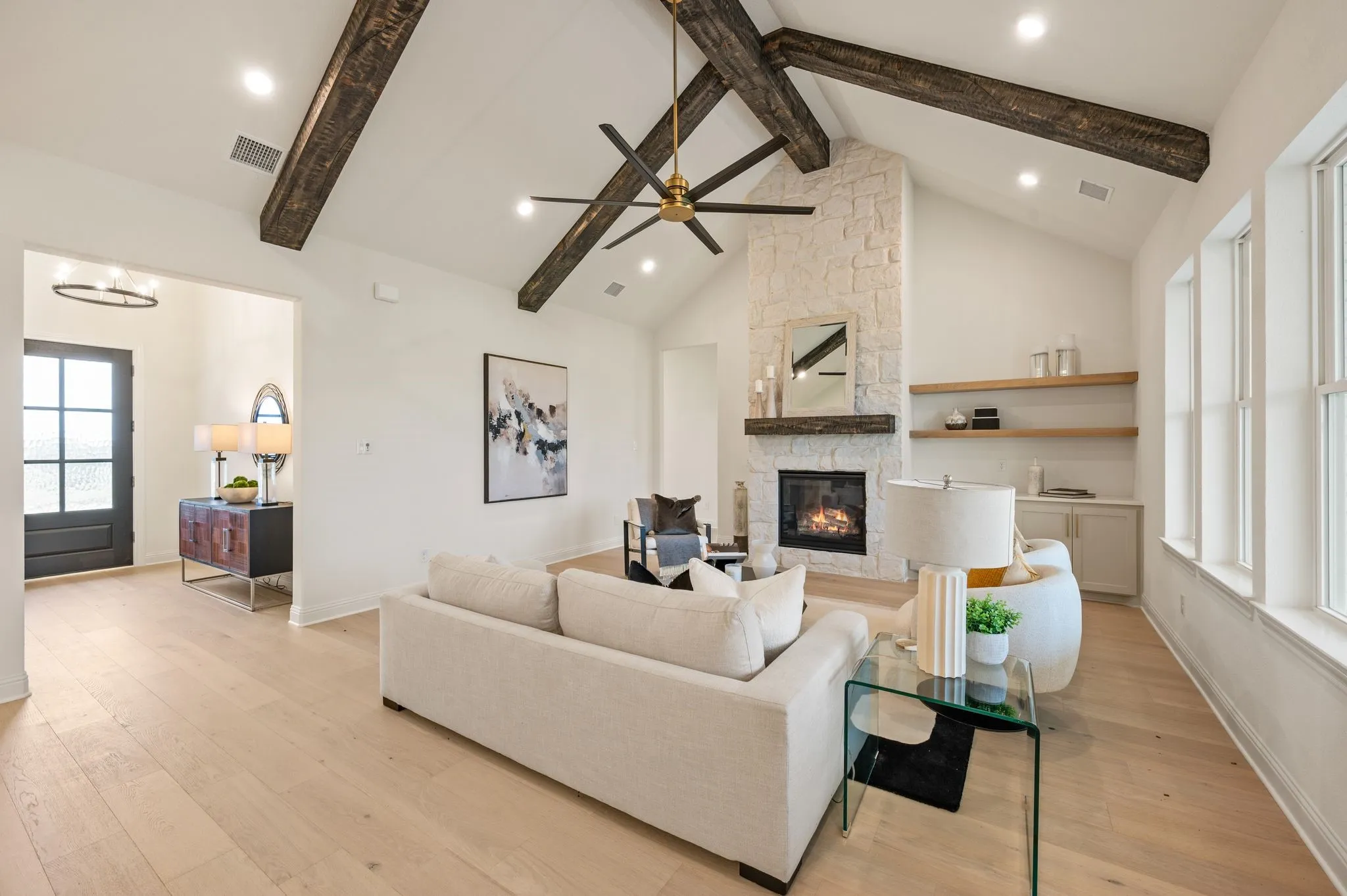 Vaulted ceilings with exposed wood beams, a stone-clad fireplace with wood mantle, light wood-finish flooring, multiple windows, and open shelves