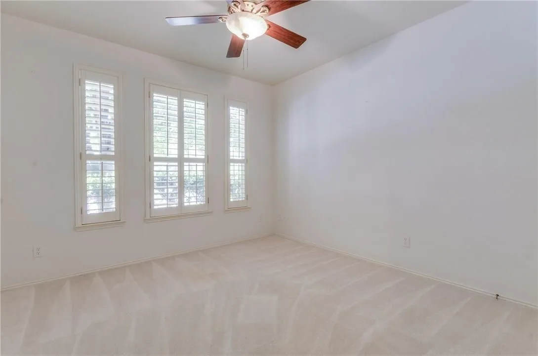 Bright interior room featuring three tall windows with louvered shutters, neutral carpeting, and a ceiling fan with light fixture