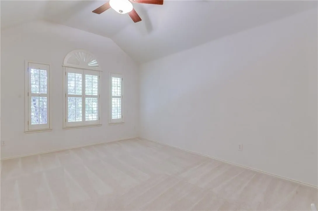 Vaulted ceiling room featuring three windows with plantation shutters, arched transom window, ceiling fan with light fixture, and neutral carpet flooring