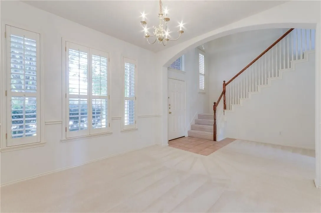 Bright interior space featuring multiple windows with plantation shutters, a chandelier, a staircase with wood newel posts and handrail, and an arched entry