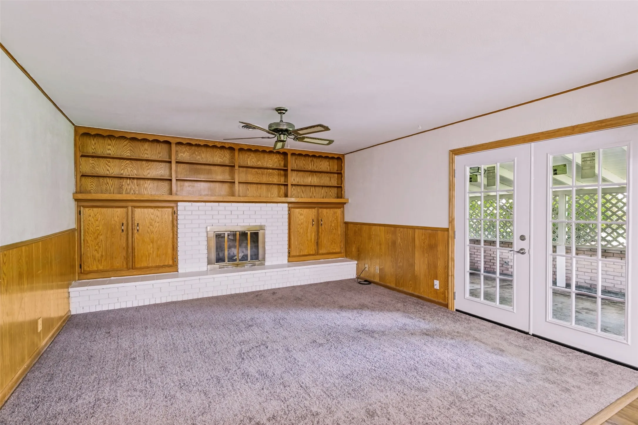 Living area featuring a white brick fireplace with a brass firebox, built-in wood shelving and cabinetry, wood wainscoting, and double French doors with grid-patterned glass