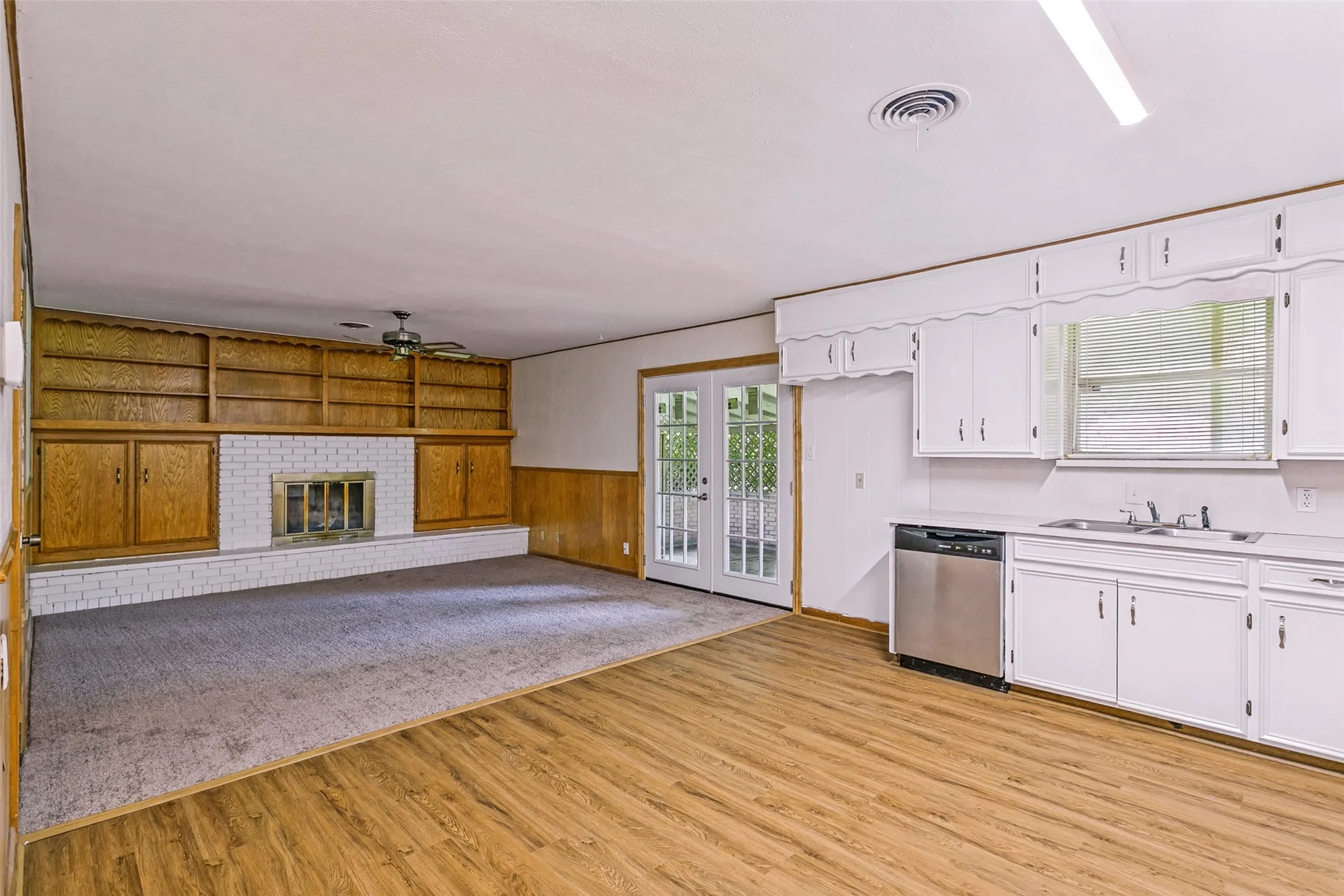 Open-concept living area featuring wood-finish flooring, white cabinetry, stainless steel dishwasher, and a built-in fireplace with white brick surround and wood shelving