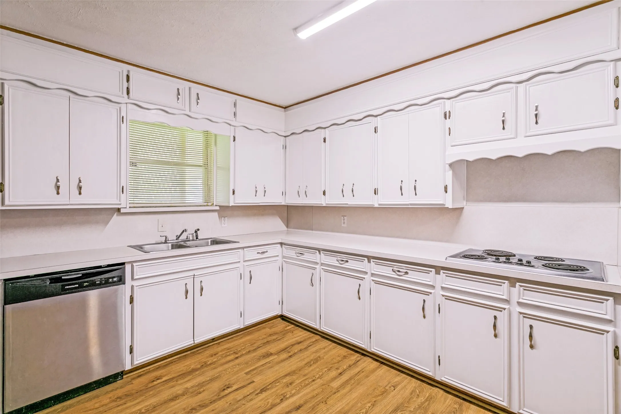 Kitchen featuring wood-finish flooring, white cabinetry with decorative trim, a stainless steel dishwasher, an integrated cooktop, and a double basin sink