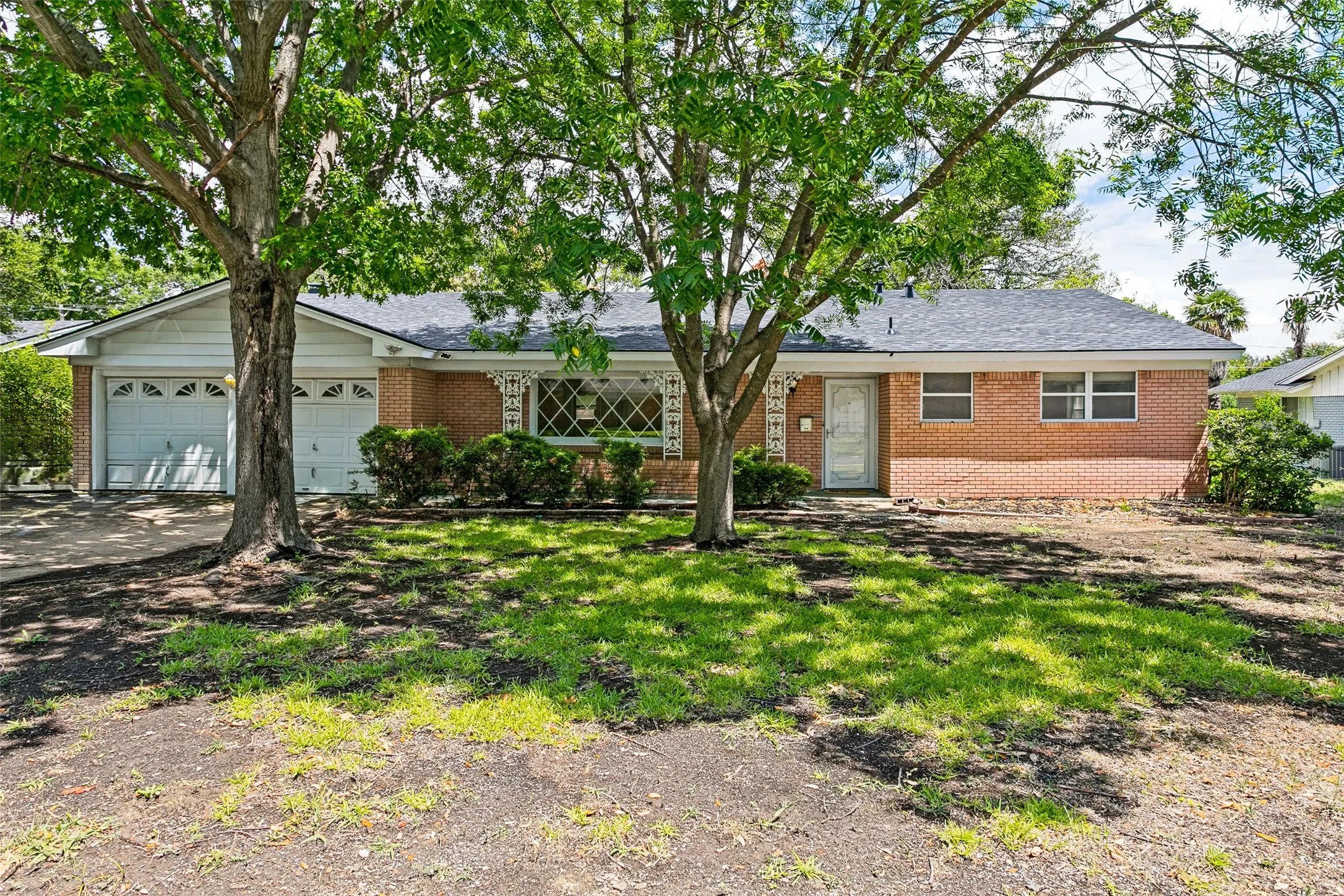 Brick facade residence featuring a two-car garage, mature front yard trees, and a decorative security door