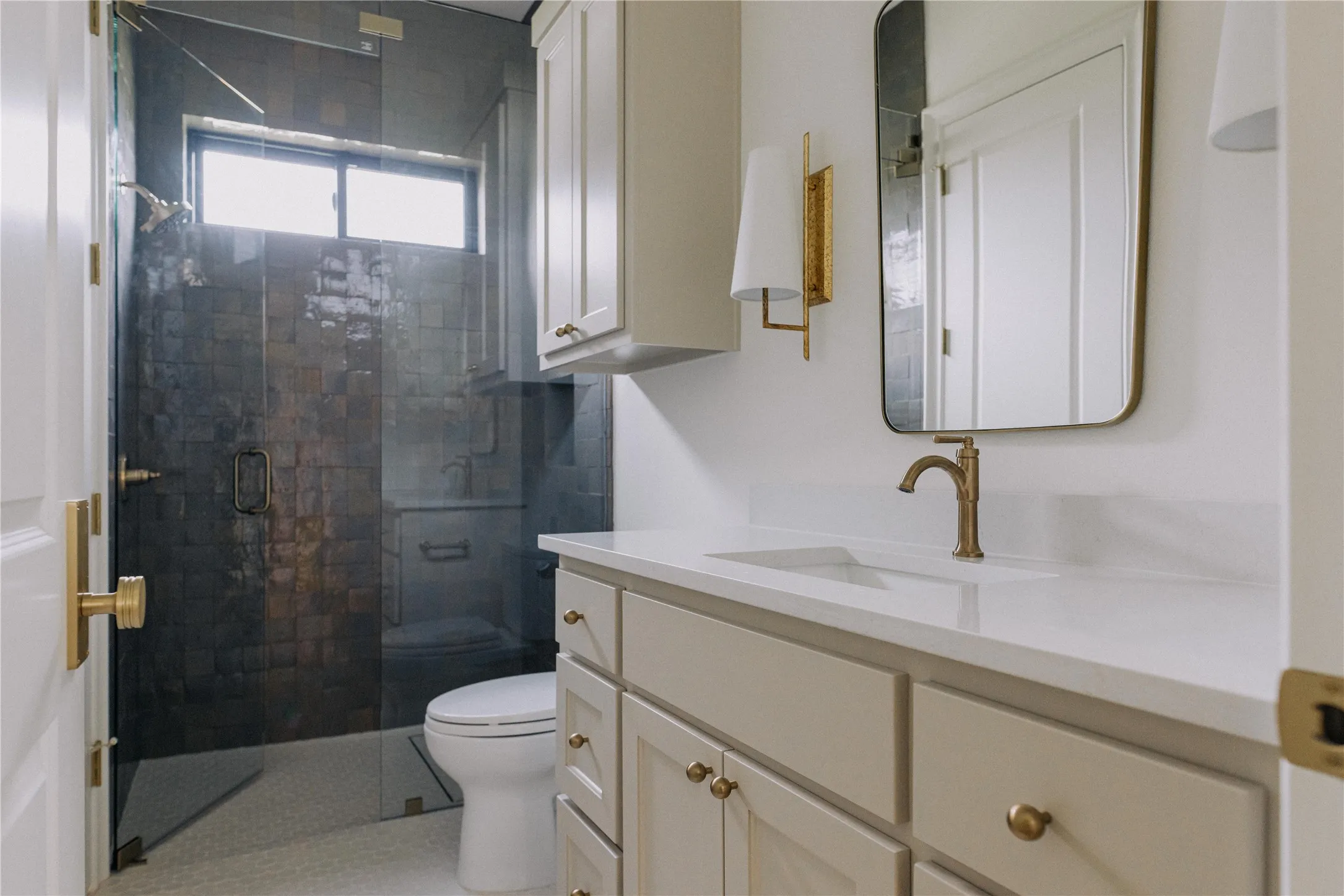 Spa-inspired primary bathroom featuring a frameless glass-enclosed shower with black tile surround, dual-sink vanity with soft-close cabinetry in neutral finish, white quartz countertop, brass hardware accents, and an arched brass-framed mirror