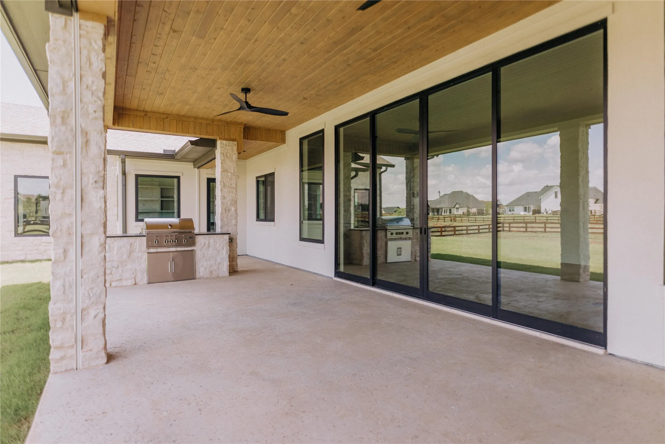 Expansive covered patio featuring an outdoor kitchen with built-in grill, stone facade, and a wood-finish ceiling with a ceiling fan