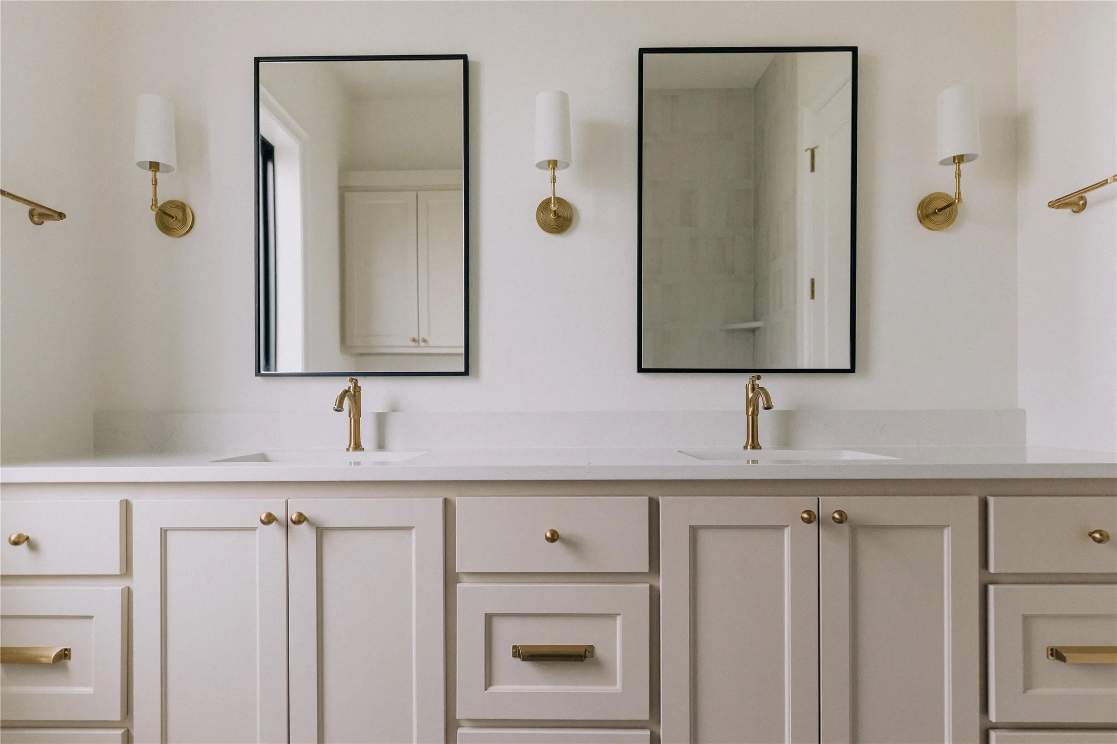 Dual-vanity bathroom featuring custom cabinetry with integrated storage, white quartz countertops, polished brass hardware and faucets, framed mirrors with black trim, and brass wall-mounted sconces flanking each sink