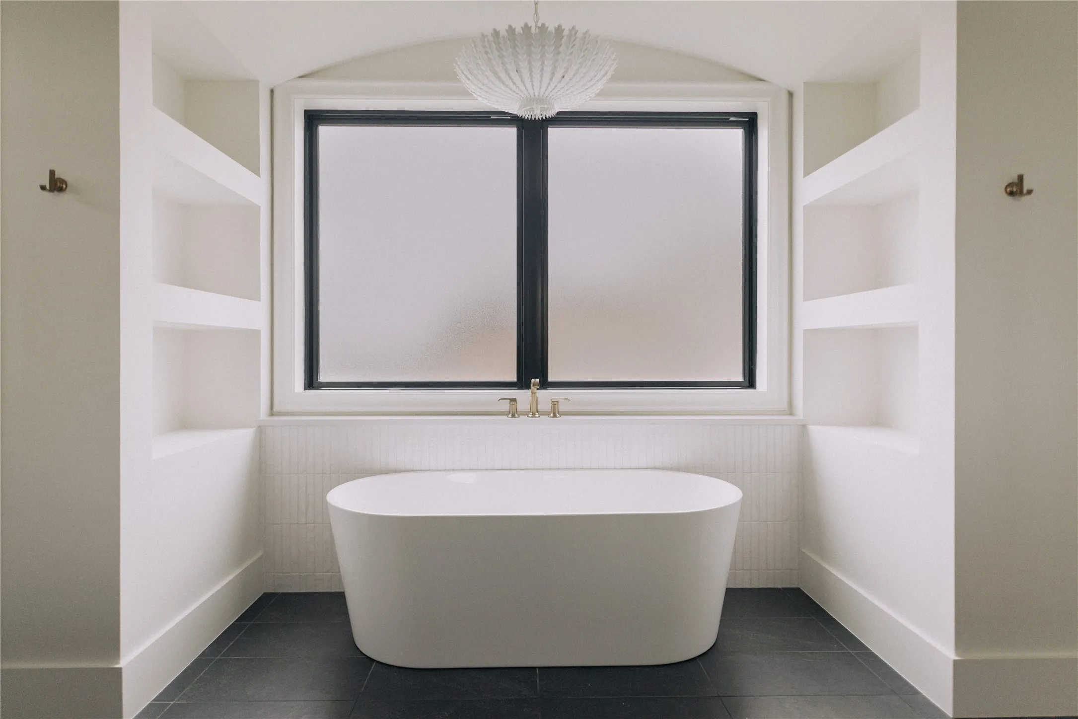 Spa-inspired primary bathroom featuring a freestanding oval soaking tub with clean-lined silhouette, positioned beneath a large dual-pane window with black aluminum framing
