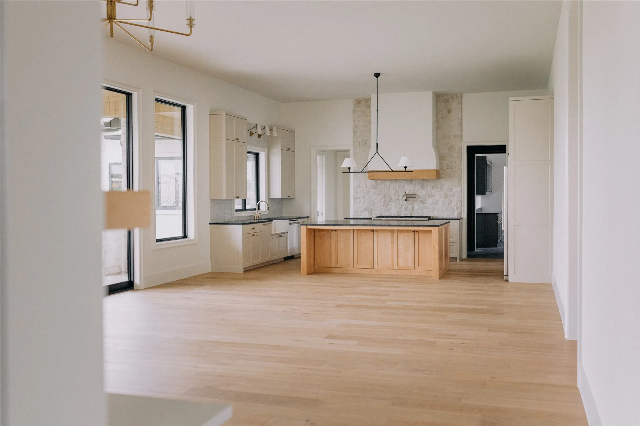 Open-concept living and kitchen featuring light wood-finish flooring throughout, custom wood cabinetry with integrated appliances, stainless steel countertops, a large center island with pendant lighting, and abundant natural light from multi-pane windows and glass doors