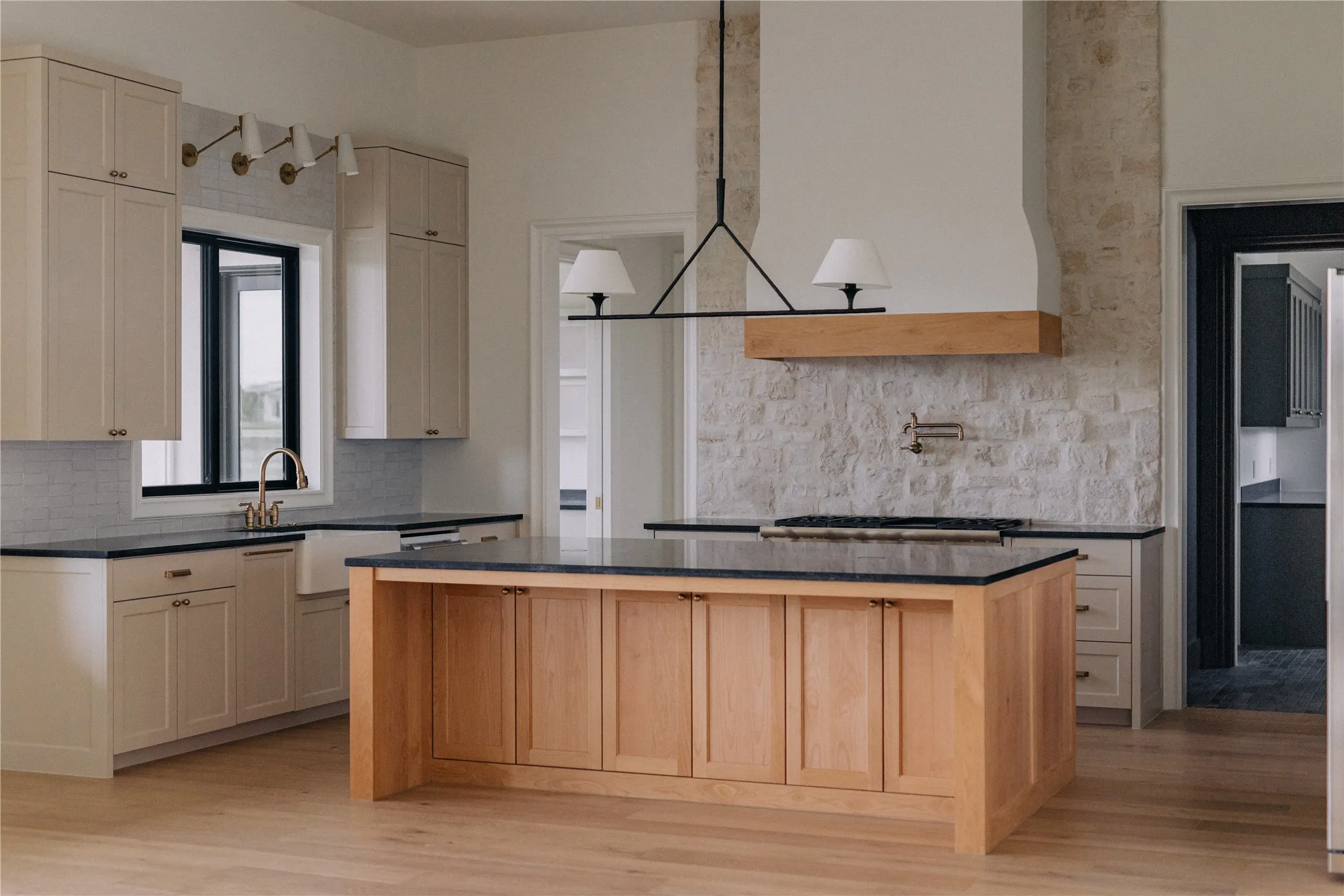 Gourmet kitchen featuring a substantial wood-finish island with black countertop, professional-grade gas cooktop, custom cabinetry in natural wood tones, stainless steel appliances, marble-look backsplash, and a statement pendant light fixture suspended from a wood beam