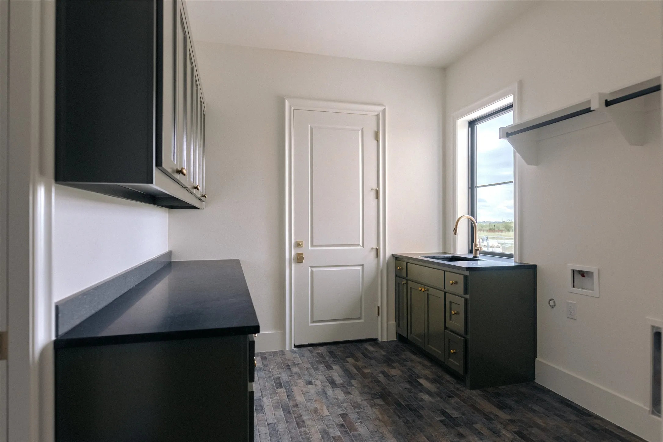 Utility room featuring wood-finish flooring, dark cabinetry with gold-tone hardware, a sink with an arched gold-tone faucet, a window, and a wall-mounted shelf