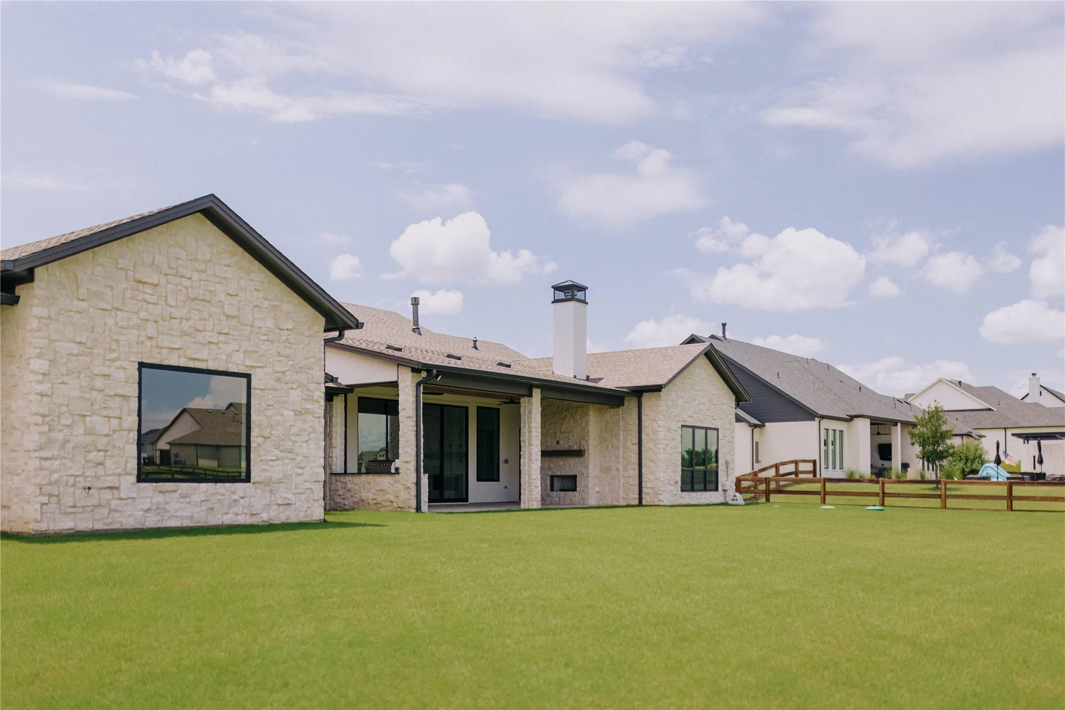 Expansive backyard featuring a manicured lawn, stone exterior with dark trim, covered patio, and a built-in outdoor fireplace