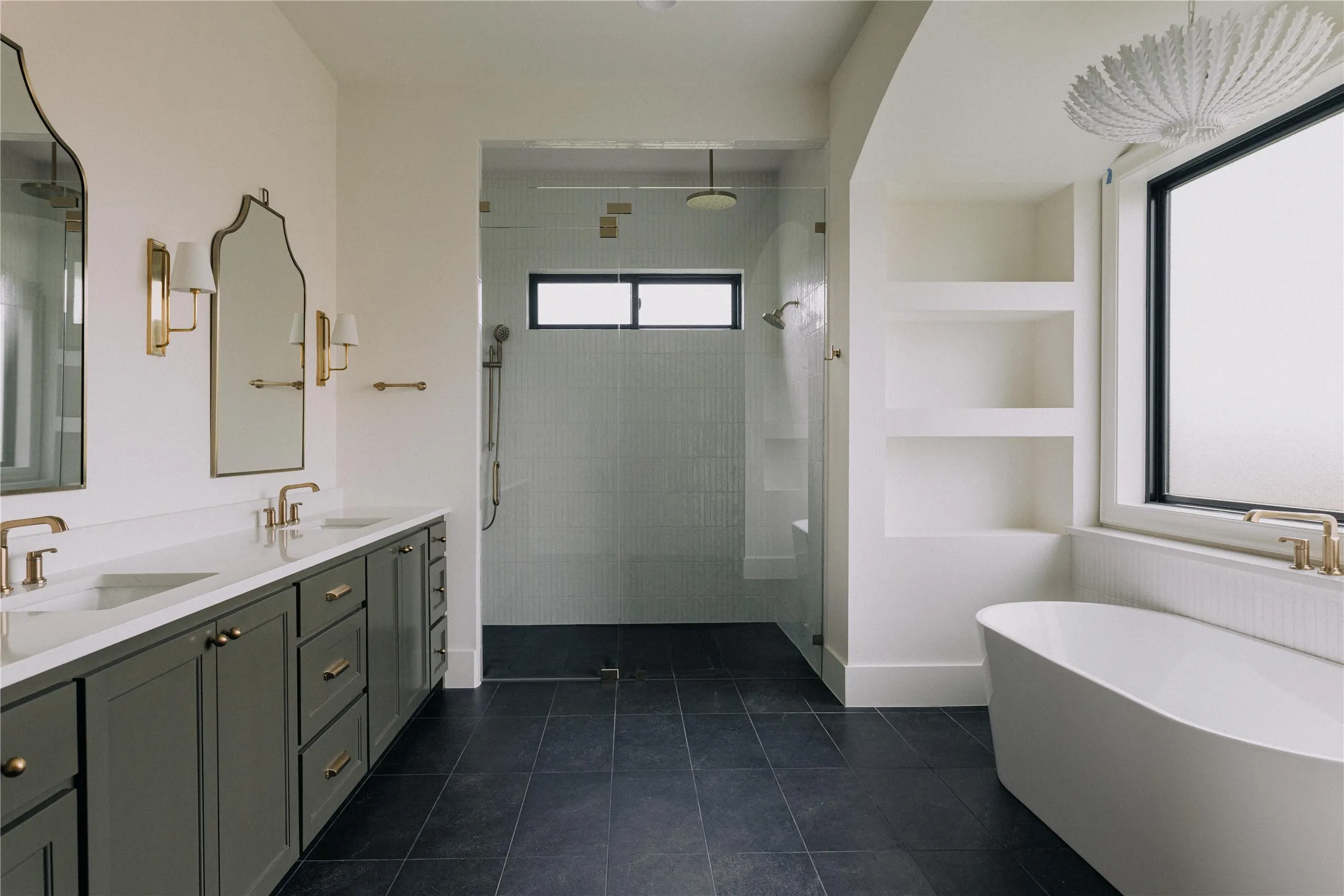 Spacious bathroom featuring a dual vanity with a white countertop and dark cabinetry, a freestanding soaking tub, a walk-in shower with clear glass enclosure, built-in shelving, and dark floor tiling