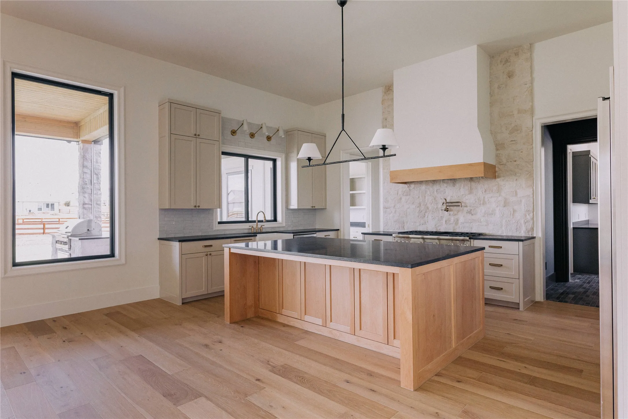Contemporary kitchen featuring a substantial wood-finish island with black countertop, custom cabinetry in soft neutral tones, stainless steel appliances, marble subway tile backsplash, wide-plank wood-finish flooring, and a modern pendant light fixture suspended above the island workspace