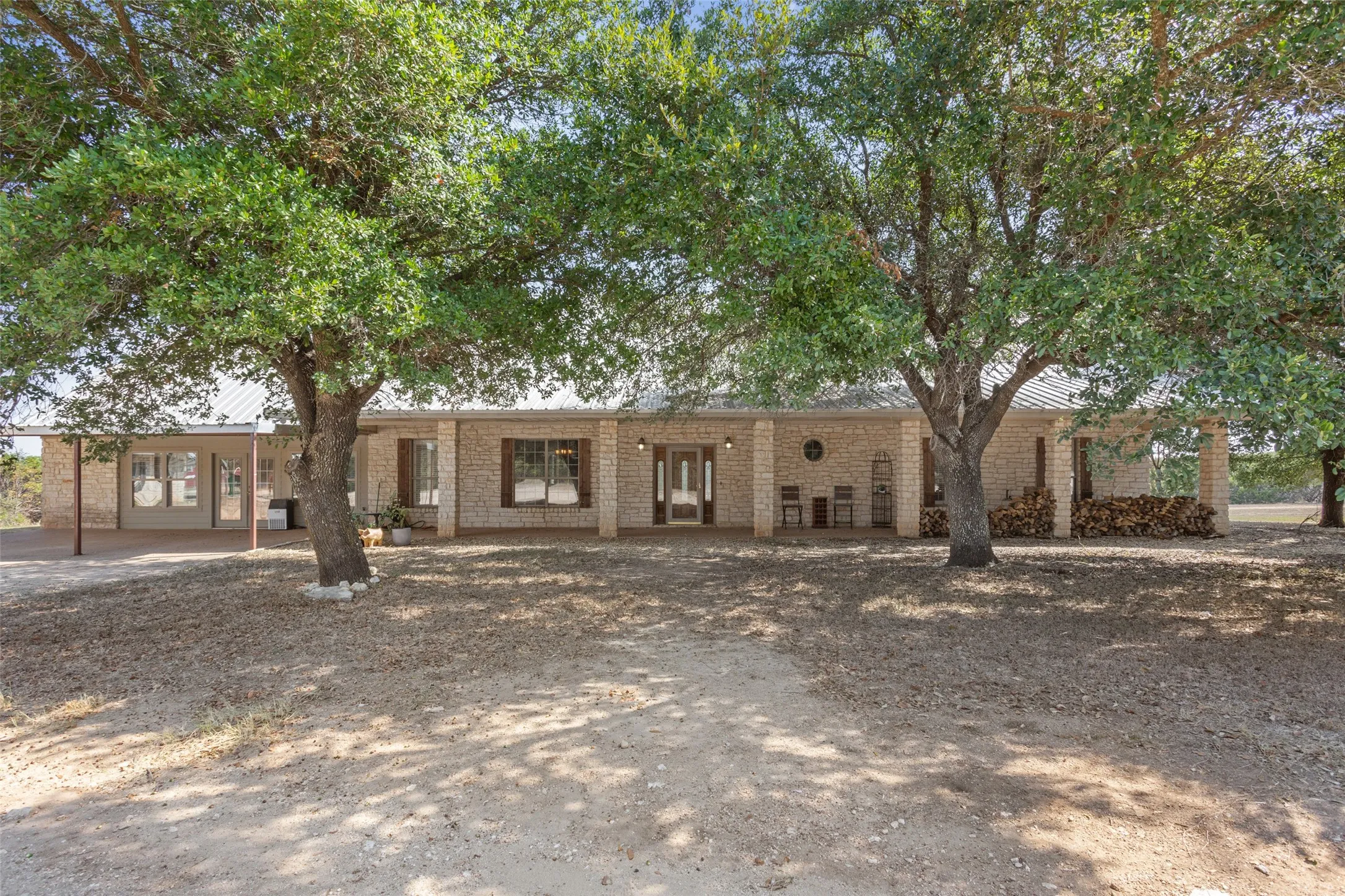 Expansive brick facade ranch-style residence featuring a metal roof, prominent mature trees, an inviting covered front porch, and substantial brick support columns
