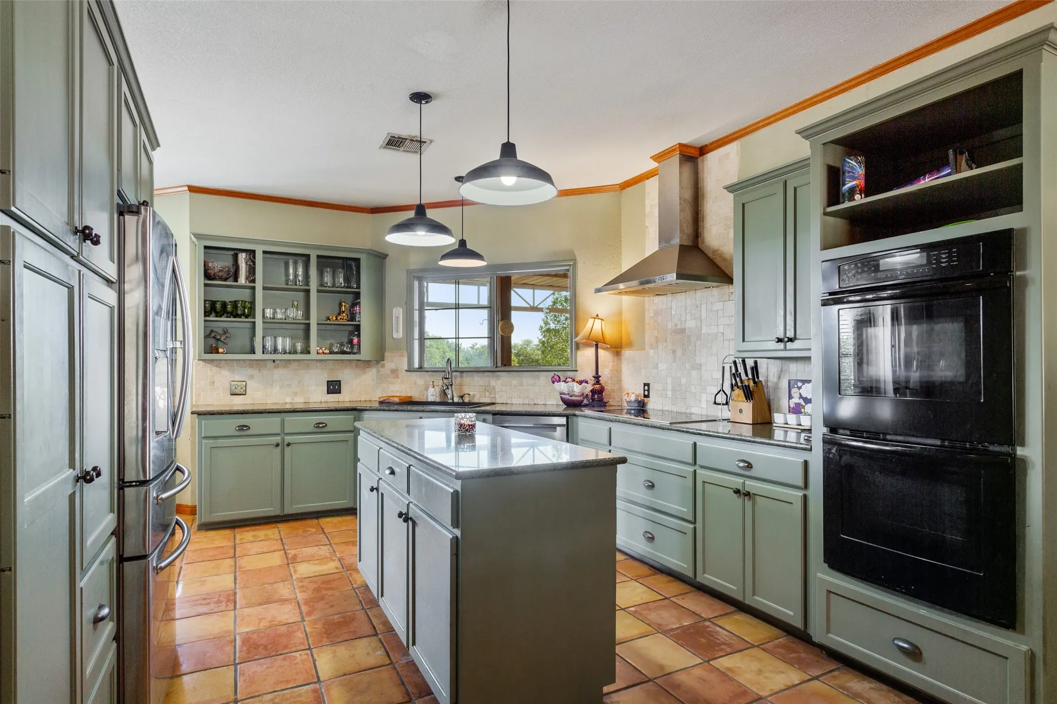 Kitchen featuring sage green cabinetry, a central island with a solid surface countertop, built-in stainless steel appliances, pendant lighting, and terracotta-style floor tiling