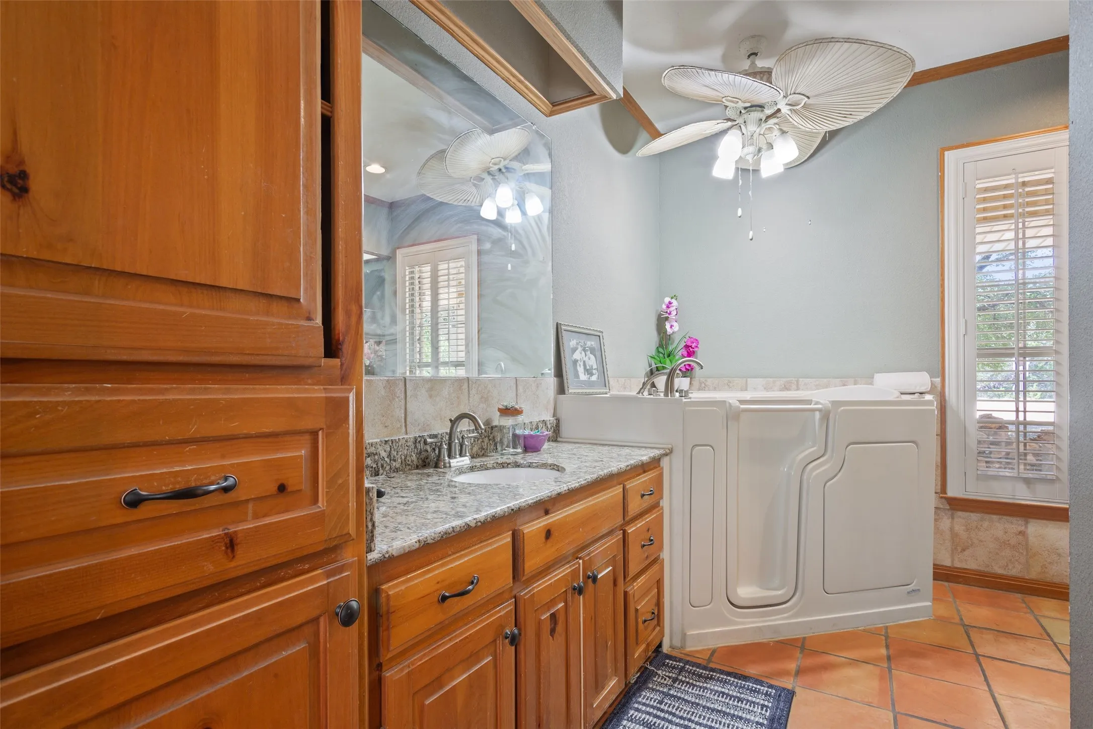 Bathroom featuring a walk-in bathtub, dual vanity with granite countertop, wood-finish cabinetry, and terracotta-style floor tiles