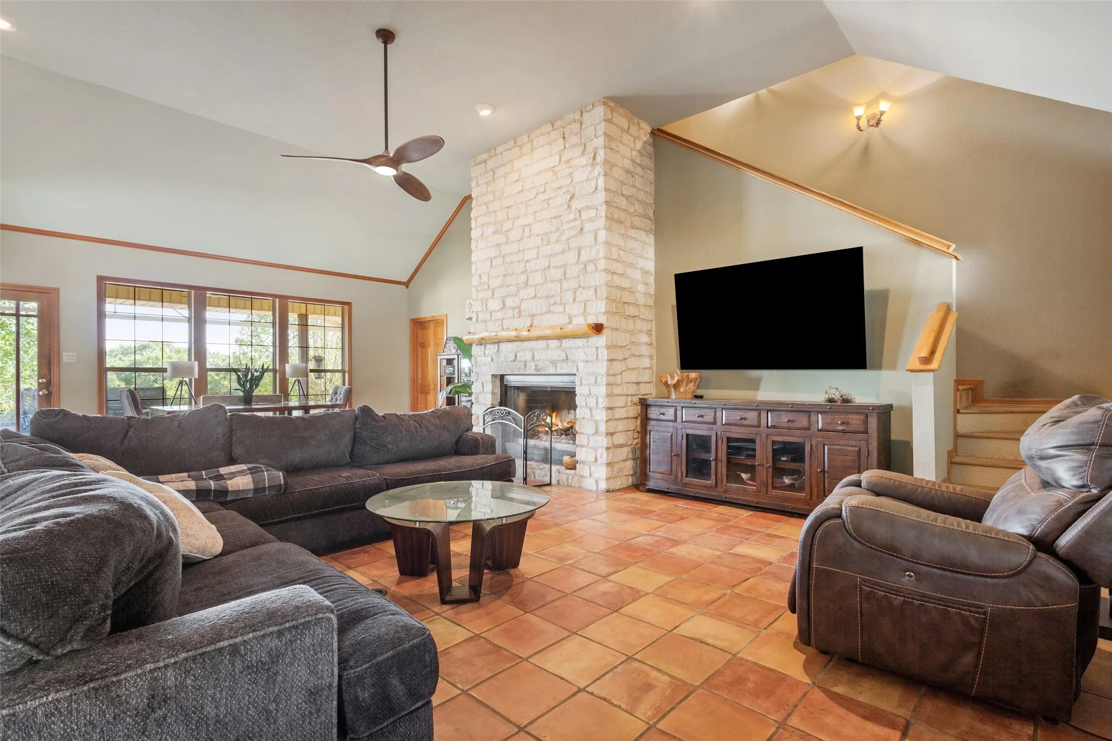 Spacious living area featuring terra cotta tile flooring, a stone-clad fireplace with a wood mantle, and a vaulted ceiling with a ceiling fan