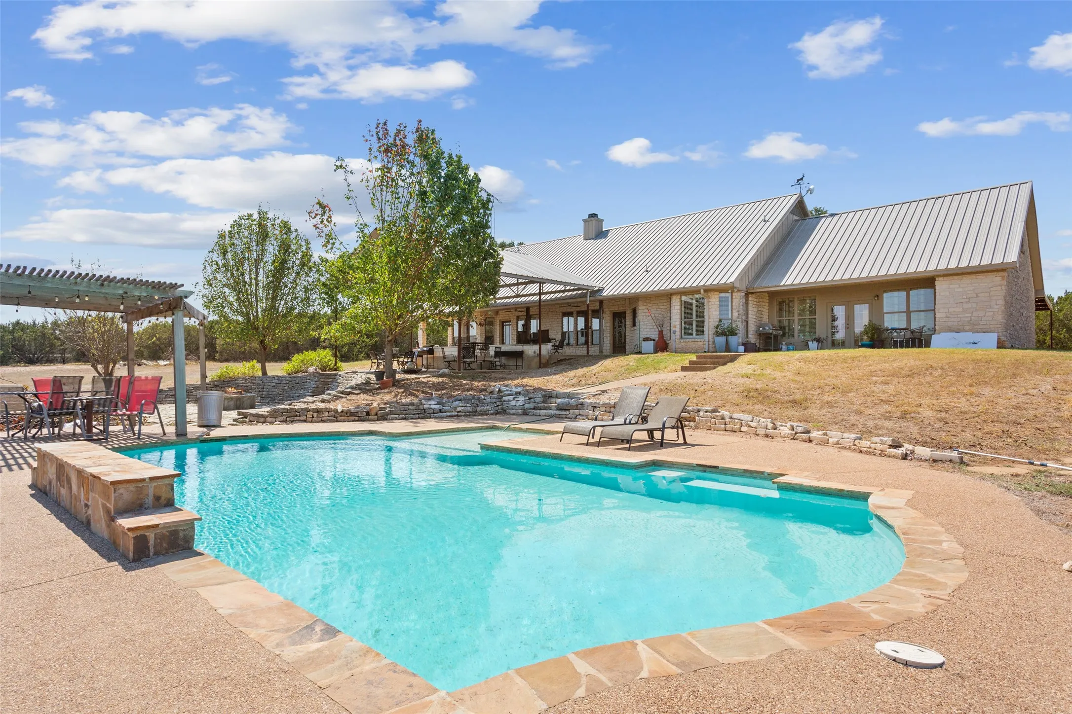 Expansive outdoor living area featuring a freeform swimming pool with stone coping, a large stone patio, and an adjacent pergola-covered dining area