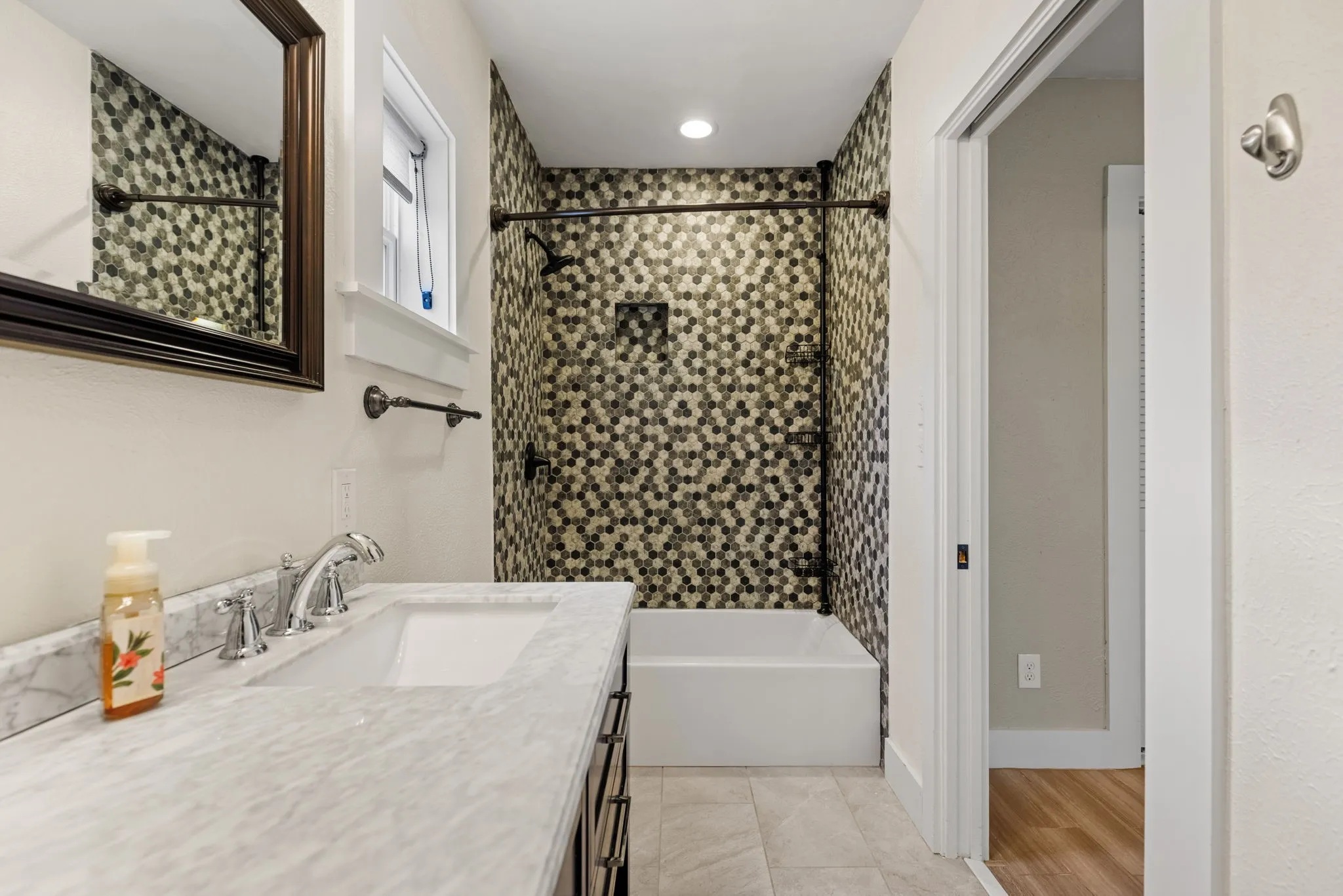 Bathroom featuring a hexagonal mosaic tile tub surround, white vanity with an integrated sink, polished chrome faucet, and a wall-mounted mirror with a dark frame